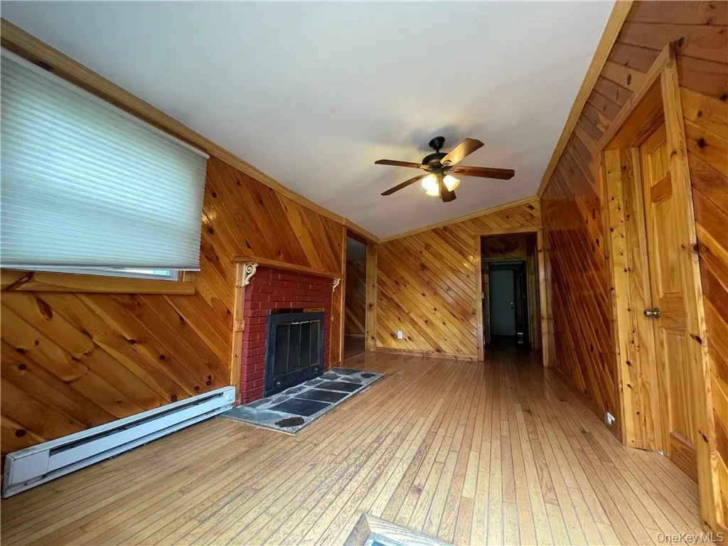 Unfurnished living room featuring a baseboard radiator, hardwood / wood-style floors, wooden walls, a fireplace with raised hearth, and crown molding Unfurnished living room featuring a baseboard radiator, hardwood / wood-style floors, wooden walls, a fireplace with raised hearth, and crown molding