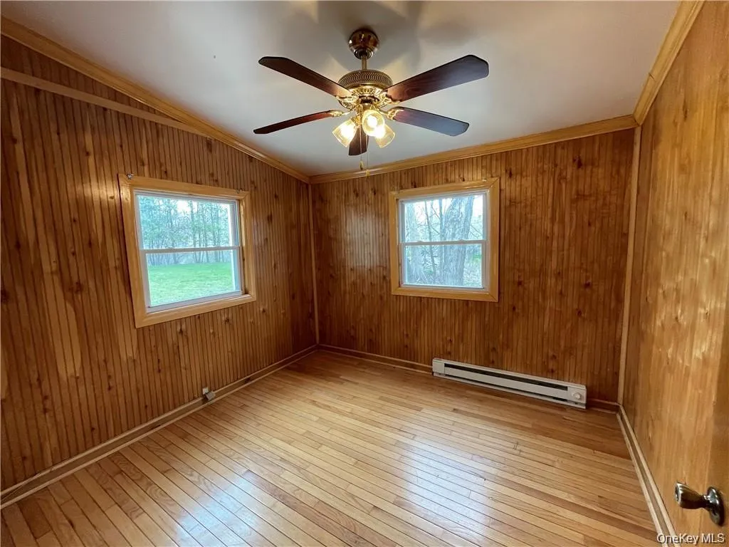 Empty room with crown molding, light wood-style flooring, a baseboard heating unit, wood walls, and ceiling fan Empty room with crown molding, light wood-style flooring, a baseboard heating unit, wood walls, and ceiling fan
