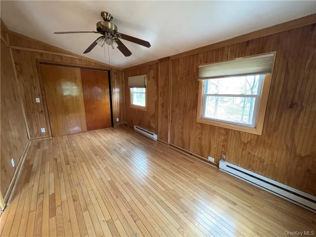 Unfurnished bedroom featuring light wood-style flooring, a closet, wooden walls, a baseboard heating unit, and ceiling fan Unfurnished bedroom featuring light wood-style flooring, a closet, wooden walls, a baseboard heating unit, and ceiling fan