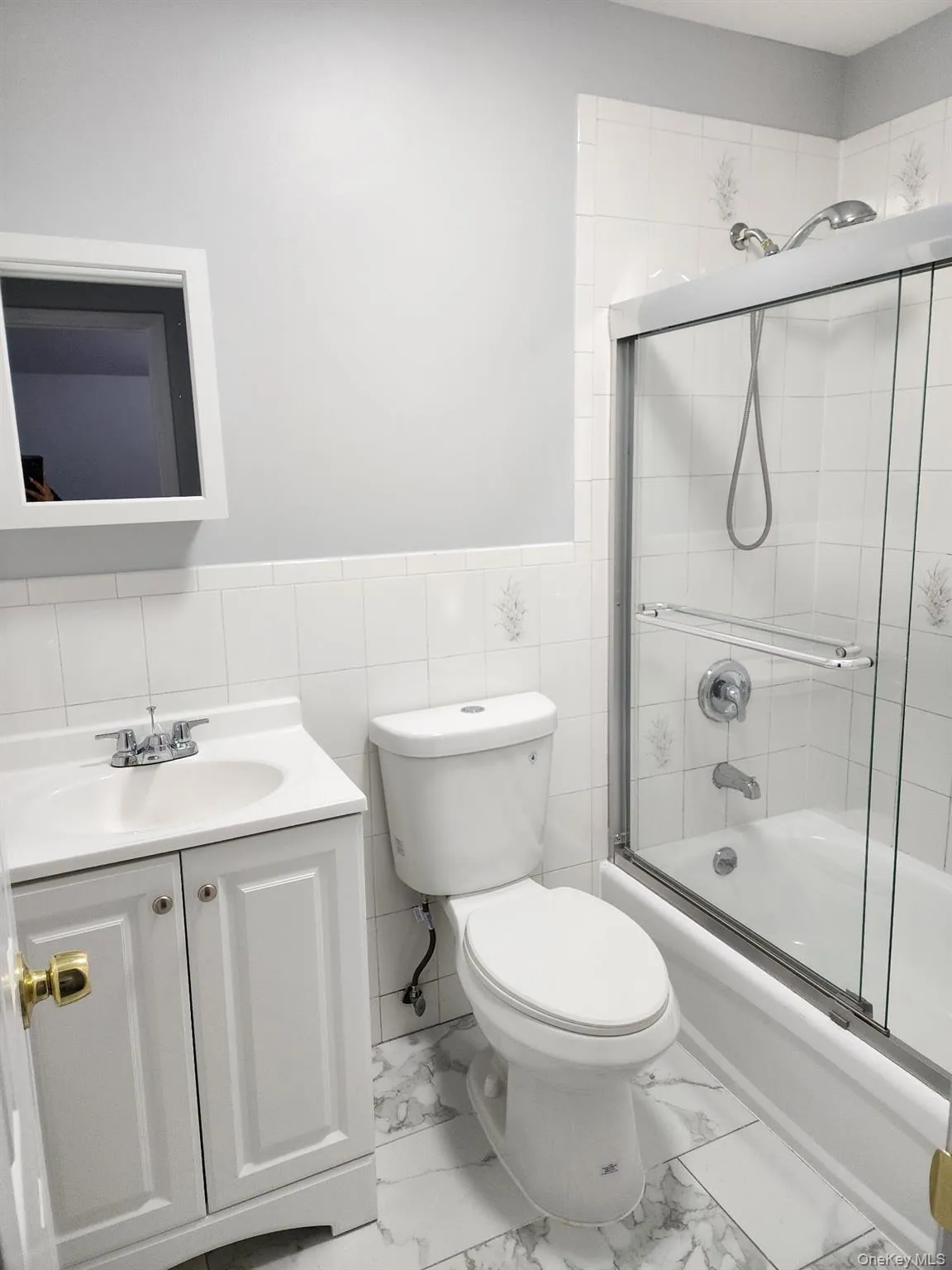 Bathroom with vanity, shower / bath combination with glass door, wainscoting, light marble finish flooring, and tile walls Bathroom with vanity, shower / bath combination with glass door, wainscoting, light marble finish flooring, and tile walls