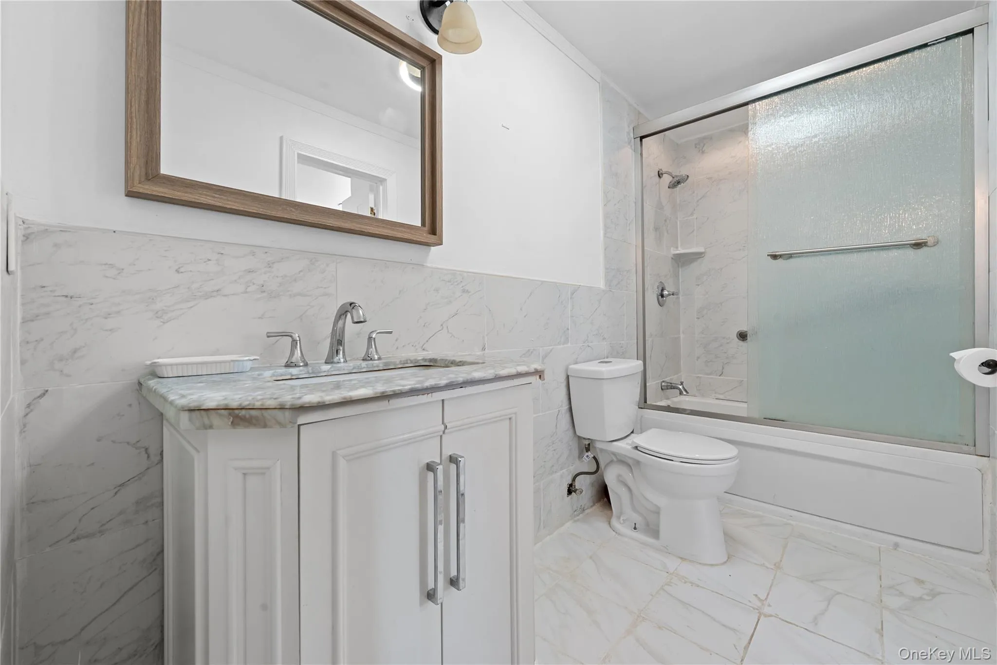 Bathroom featuring tile walls, vanity, enclosed tub / shower combo, wainscoting, and light marble finish floors Bathroom featuring tile walls, vanity, enclosed tub / shower combo, wainscoting, and light marble finish floors
