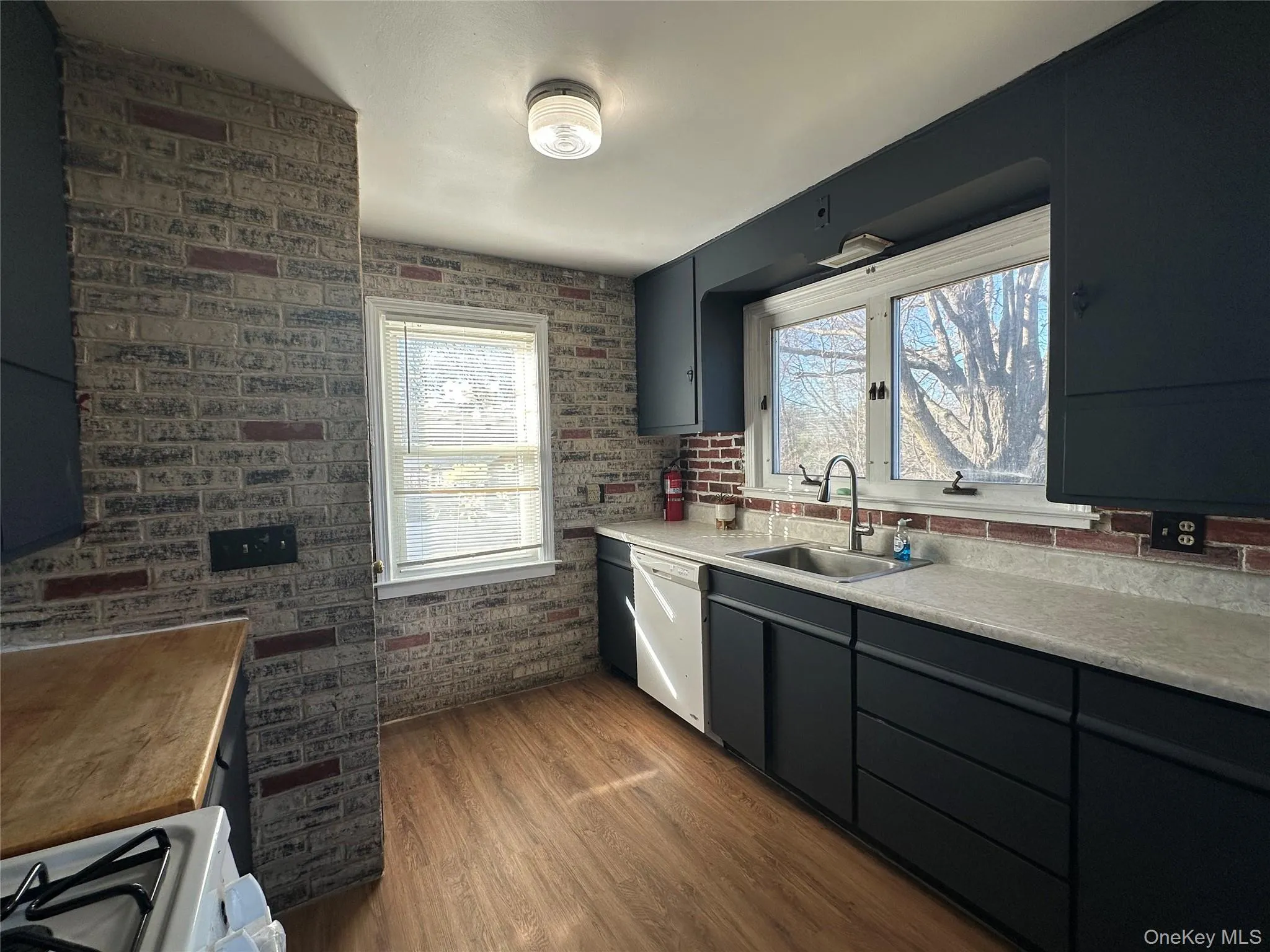 Kitchen with dark wood finished floors, light countertops, brick wall, white appliances, and gray cabinetry Kitchen with dark wood finished floors, light countertops, brick wall, white appliances, and gray cabinetry