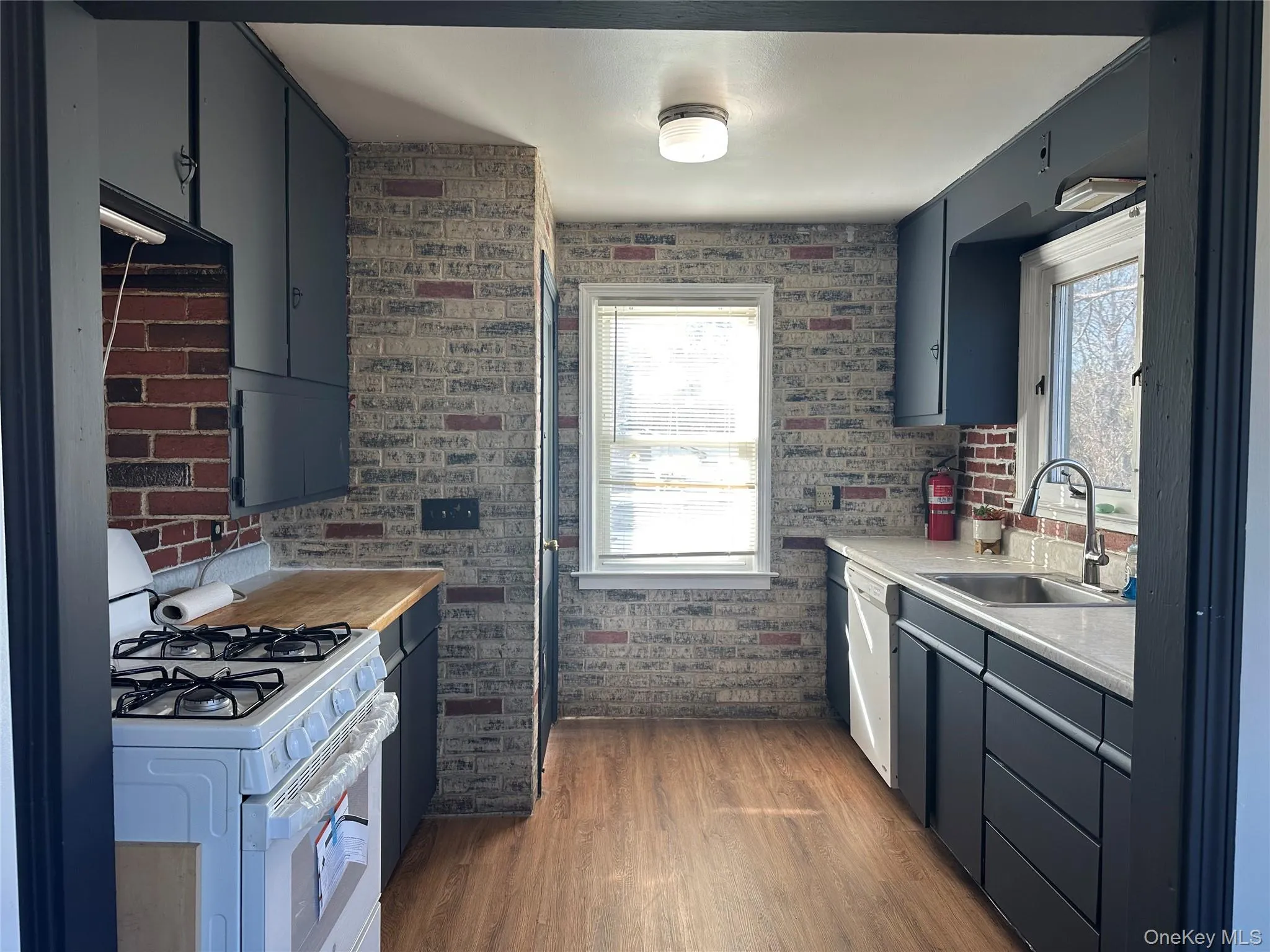 Kitchen with white appliances, dark wood-style flooring, blue cabinets, light countertops, and brick wall Kitchen with white appliances, dark wood-style flooring, blue cabinets, light countertops, and brick wall