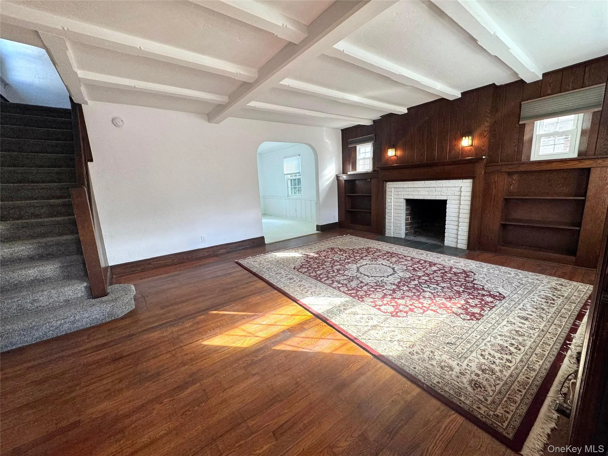 Living room featuring dark wood-style floors, beam ceiling, a brick fireplace, arched walkways, and built in features Living room featuring dark wood-style floors, beam ceiling, a brick fireplace, arched walkways, and built in features