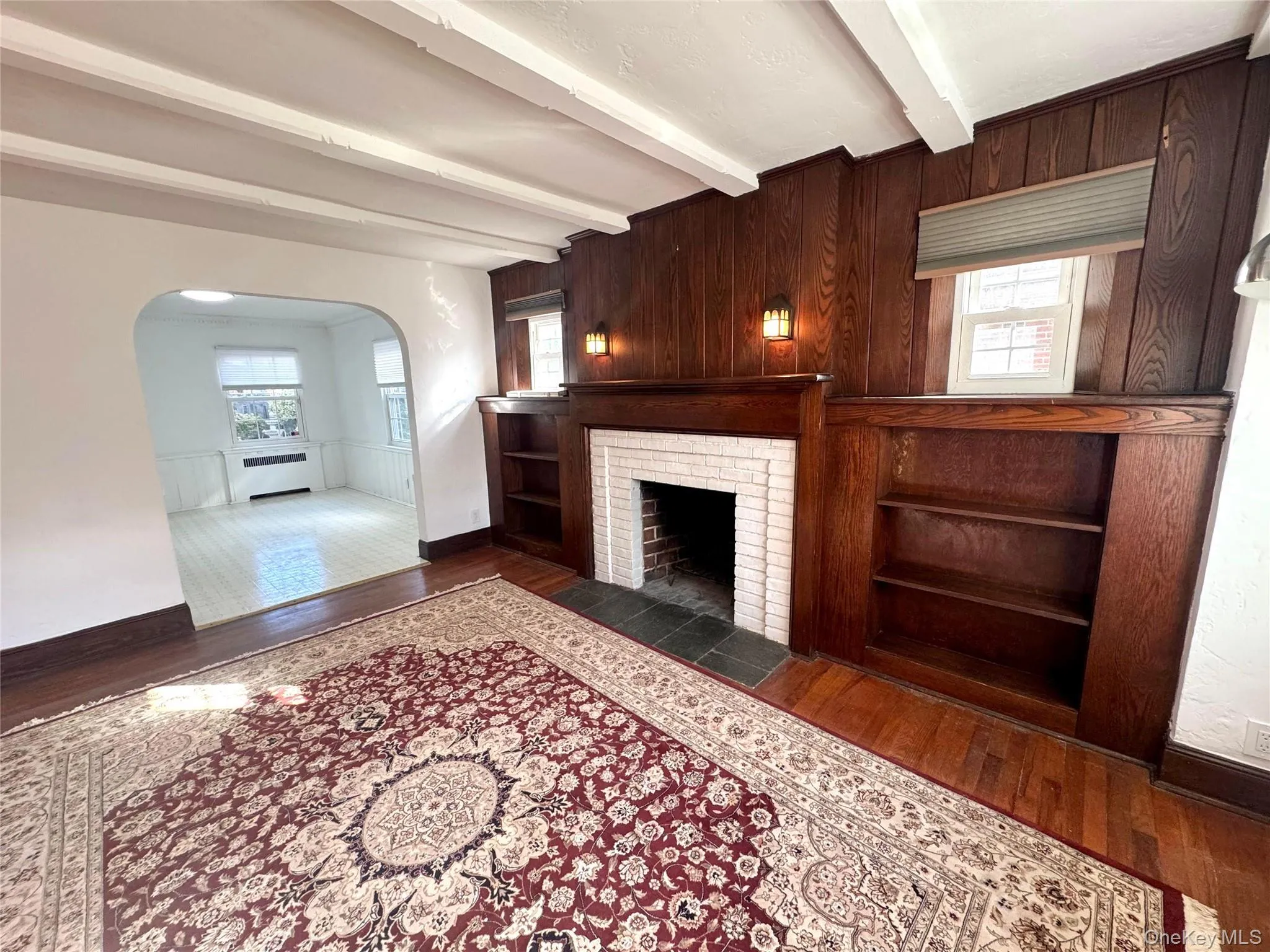 Unfurnished living room with dark wood-style flooring, a brick fireplace, arched walkways, beamed ceiling, and wood walls Unfurnished living room with dark wood-style flooring, a brick fireplace, arched walkways, beamed ceiling, and wood walls