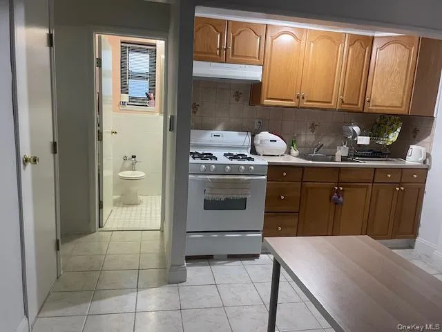 Kitchen with light countertops, white range with gas stovetop, brown cabinetry, under cabinet range hood, and backsplash Kitchen with light countertops, white range with gas stovetop, brown cabinetry, under cabinet range hood, and backsplash