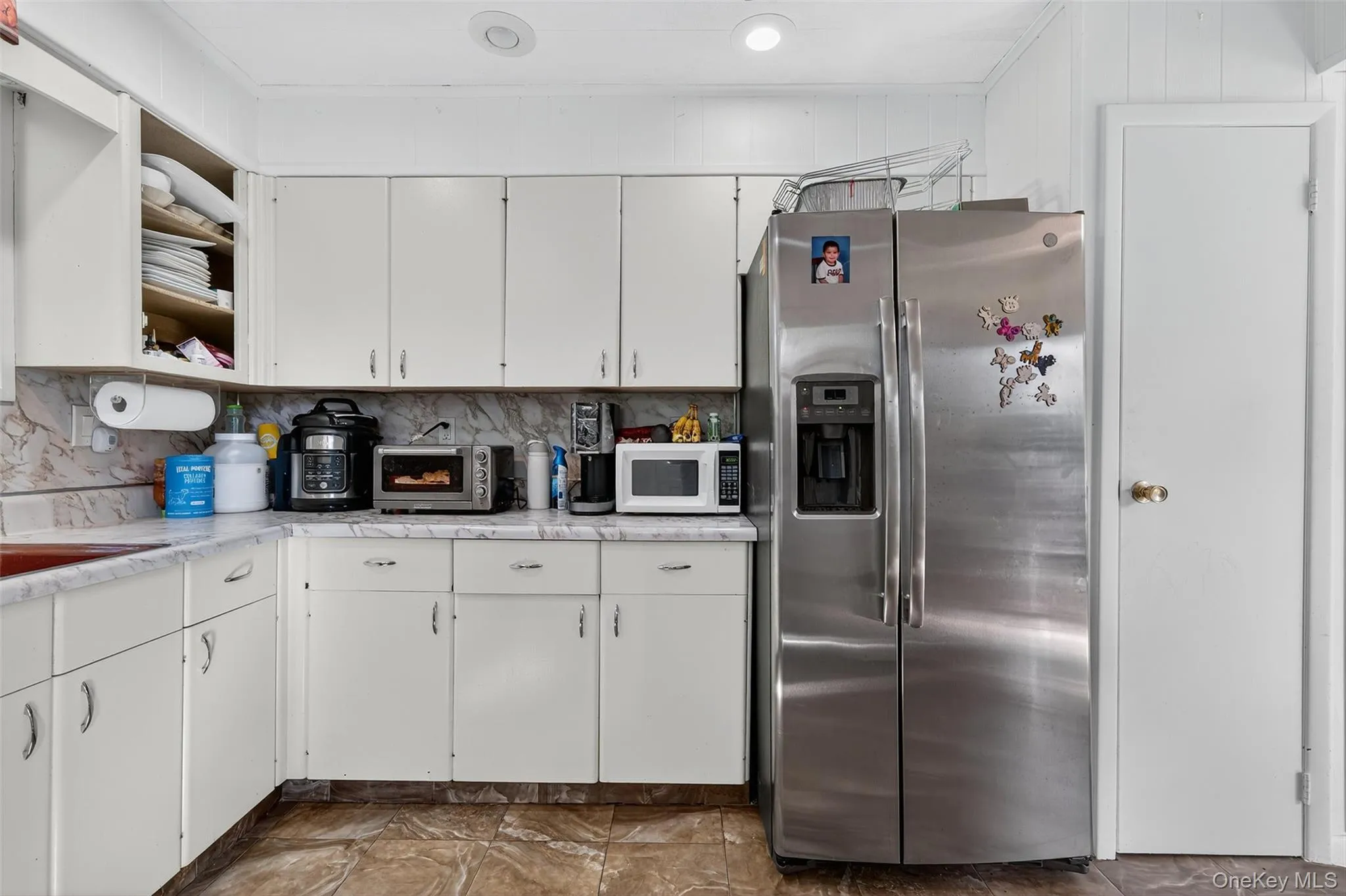Kitchen with open shelves, stainless steel fridge, white cabinets, white microwave, and tasteful backsplash Kitchen with open shelves, stainless steel fridge, white cabinets, white microwave, and tasteful backsplash