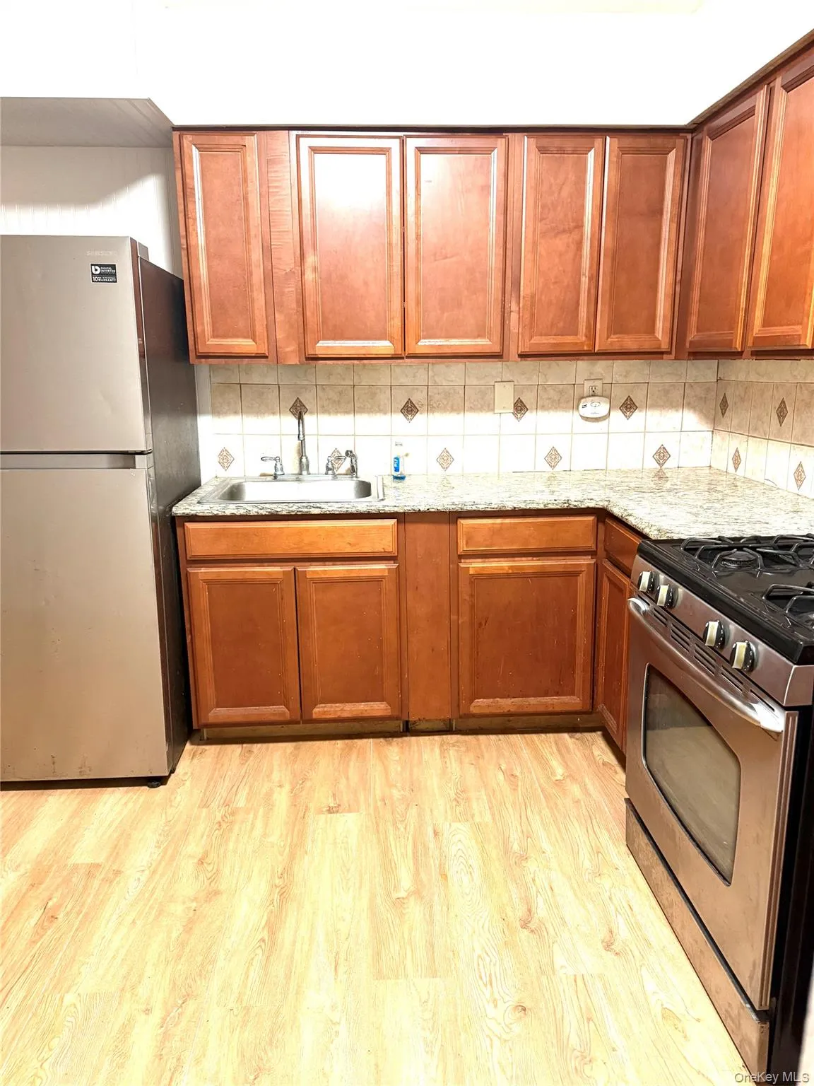 Kitchen with brown cabinetry, stainless steel appliances, light wood-style flooring, backsplash, and light stone counters Kitchen with brown cabinetry, stainless steel appliances, light wood-style flooring, backsplash, and light stone counters