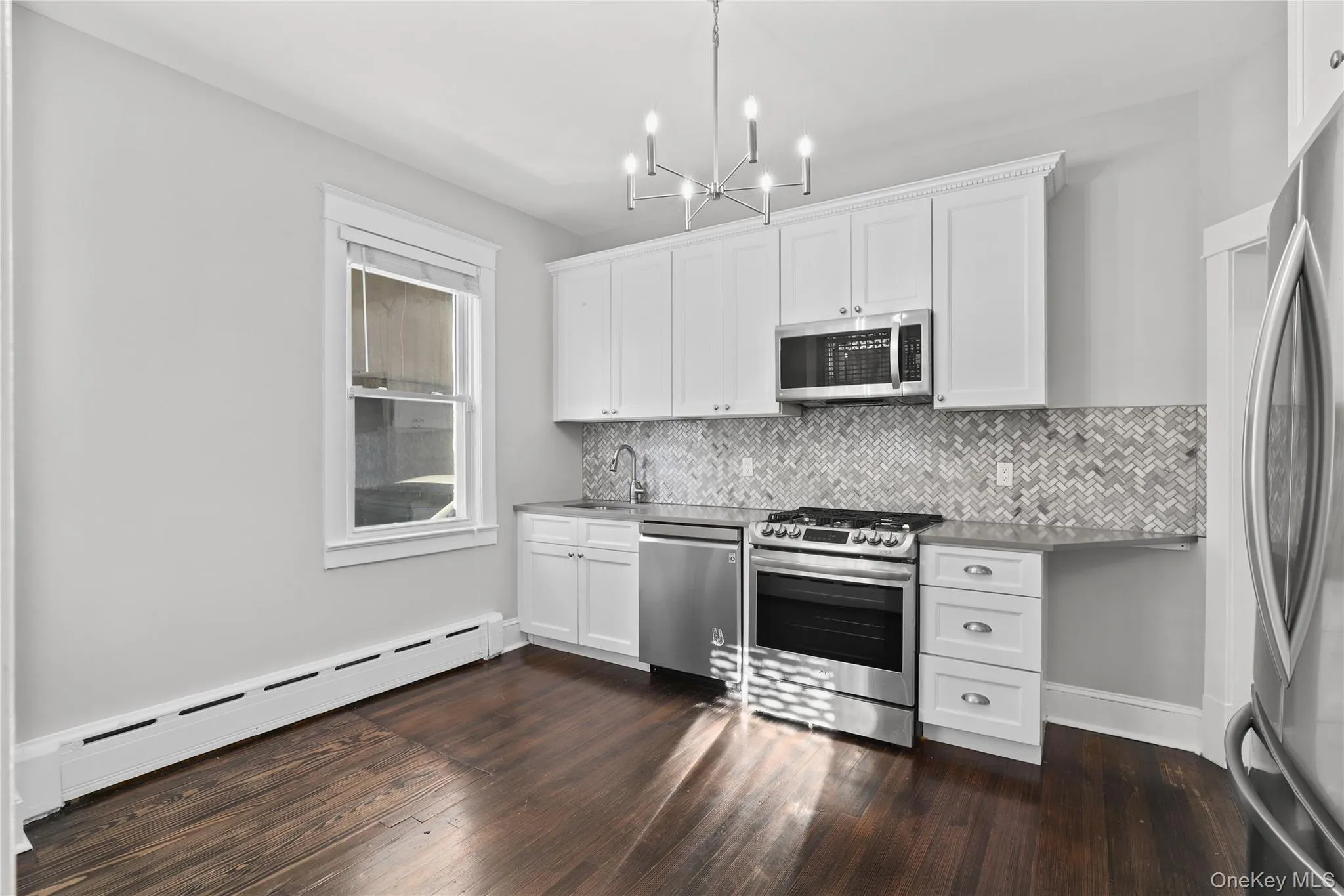 Kitchen featuring stainless steel appliances, a baseboard heating unit, white cabinets, backsplash, and a chandelier Kitchen featuring stainless steel appliances, a baseboard heating unit, white cabinets, backsplash, and a chandelier