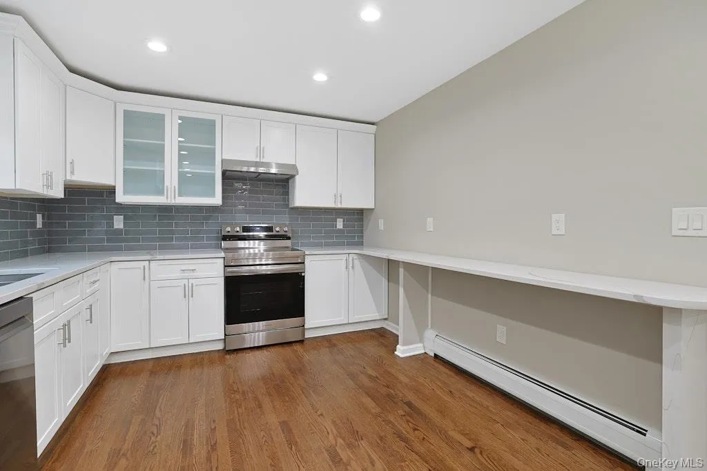 Kitchen featuring a baseboard heating unit, stainless steel appliances, tasteful backsplash, white cabinetry, and dark wood-style floors Kitchen featuring a baseboard heating unit, stainless steel appliances, tasteful backsplash, white cabinetry, and dark wood-style floors