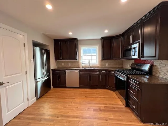 Kitchen featuring dark brown cabinets, stainless steel appliances, backsplash, light wood finished floors, and dark stone countertops Kitchen featuring dark brown cabinets, stainless steel appliances, backsplash, light wood finished floors, and dark stone countertops