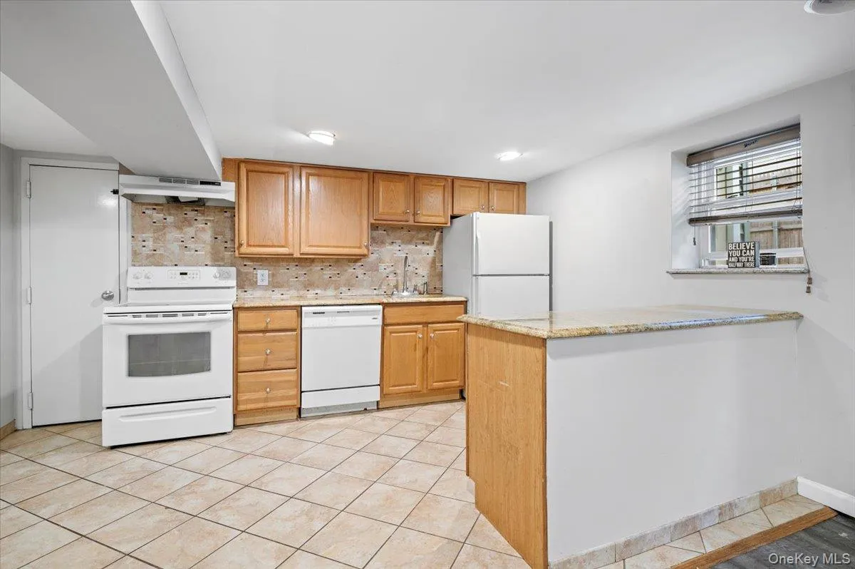 Kitchen featuring white appliances, decorative backsplash, light stone counters, light tile patterned floors, and brown cabinetry Kitchen featuring white appliances, decorative backsplash, light stone counters, light tile patterned floors, and brown cabinetry