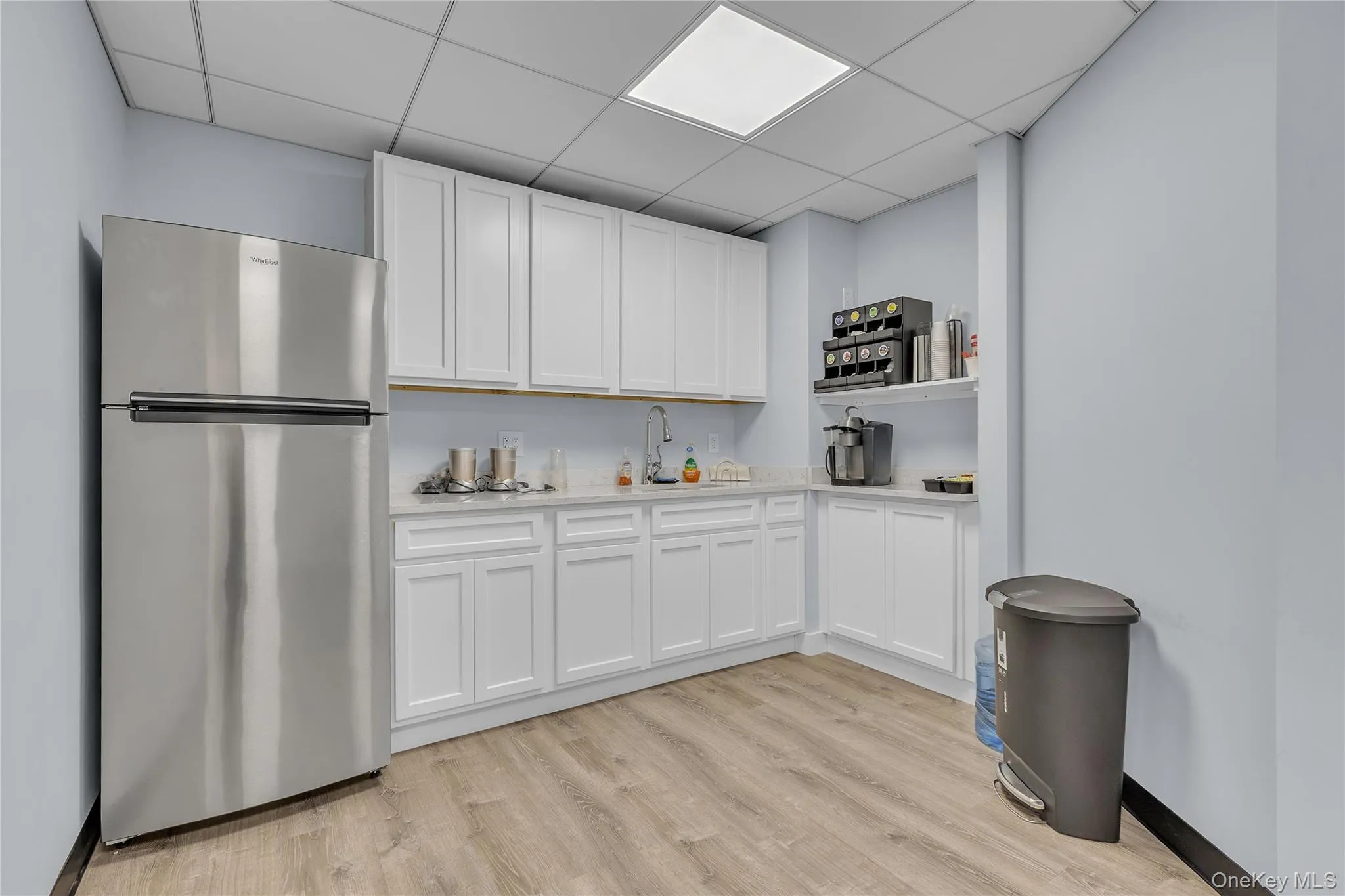 Kitchen featuring freestanding refrigerator, white cabinets, a drop ceiling, light wood-style flooring, and light stone counters Kitchen featuring freestanding refrigerator, white cabinets, a drop ceiling, light wood-style flooring, and light stone counters