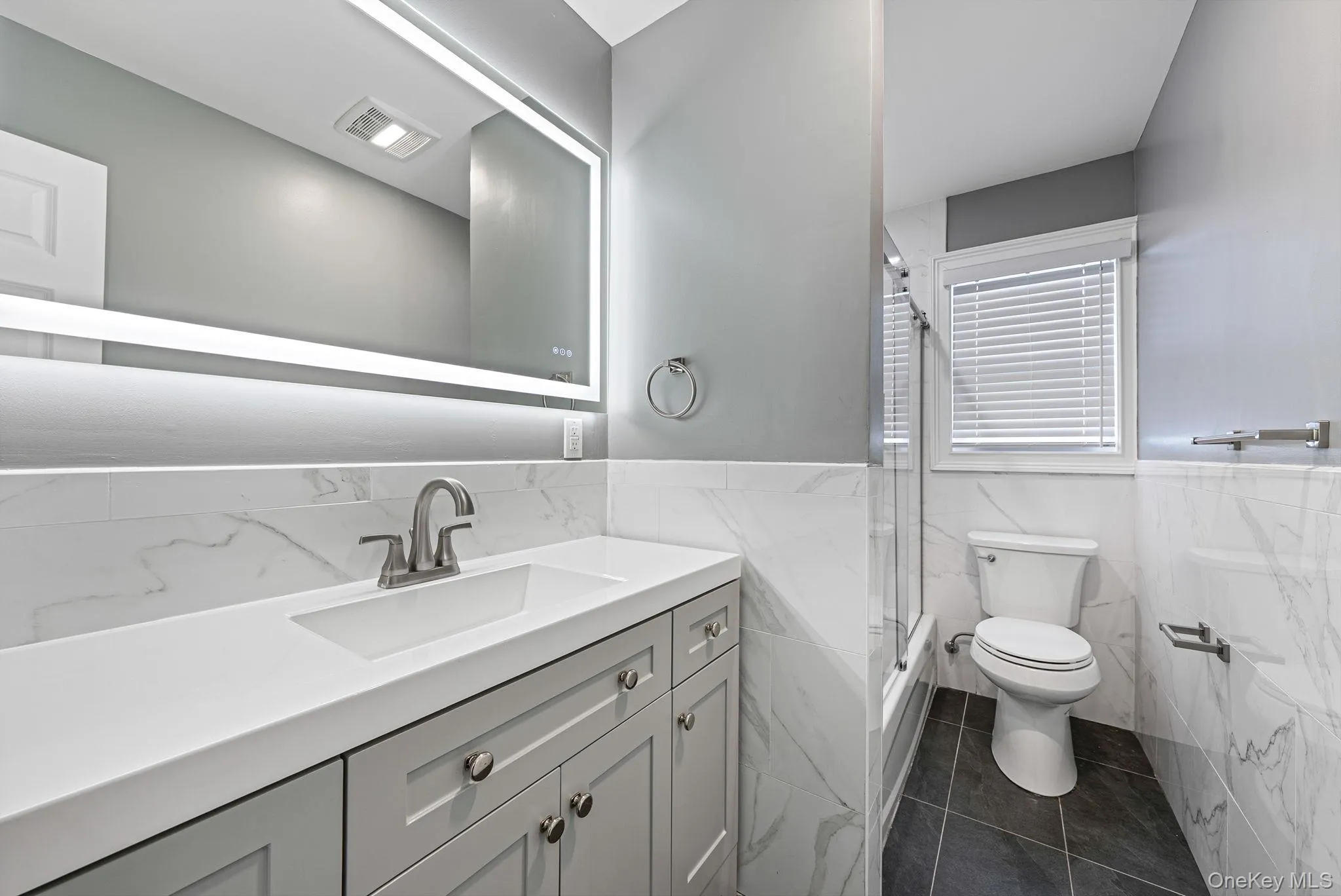 Full bathroom featuring vanity, shower / bath combination with glass door, a wainscoted wall, dark tile patterned flooring, and tile walls Full bathroom featuring vanity, shower / bath combination with glass door, a wainscoted wall, dark tile patterned flooring, and tile walls