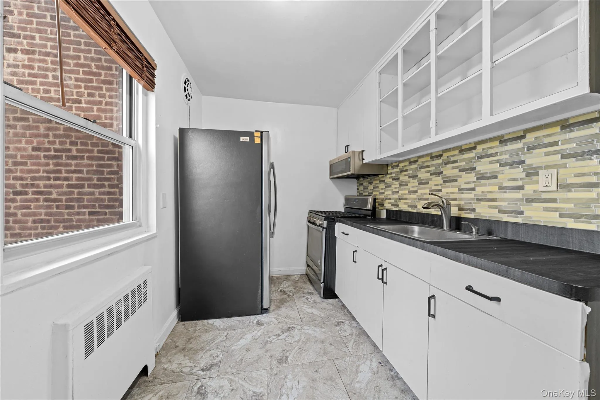 Kitchen featuring stainless steel appliances, radiator heating unit, dark countertops, open shelves, and white cabinets Kitchen featuring stainless steel appliances, radiator heating unit, dark countertops, open shelves, and white cabinets