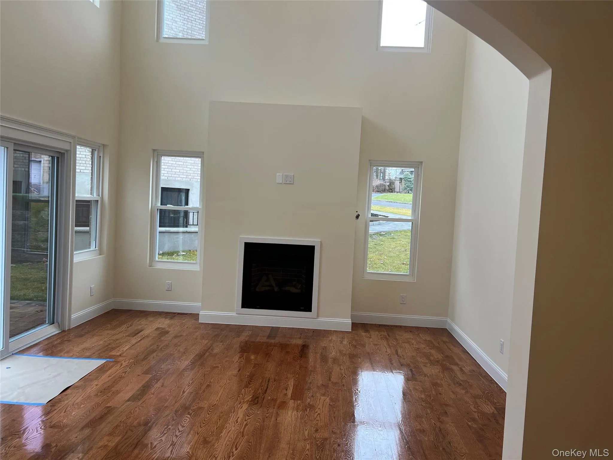Unfurnished living room with a high ceiling, dark wood-style floors, a fireplace, and healthy amount of natural light Unfurnished living room with a high ceiling, dark wood-style floors, a fireplace, and healthy amount of natural light