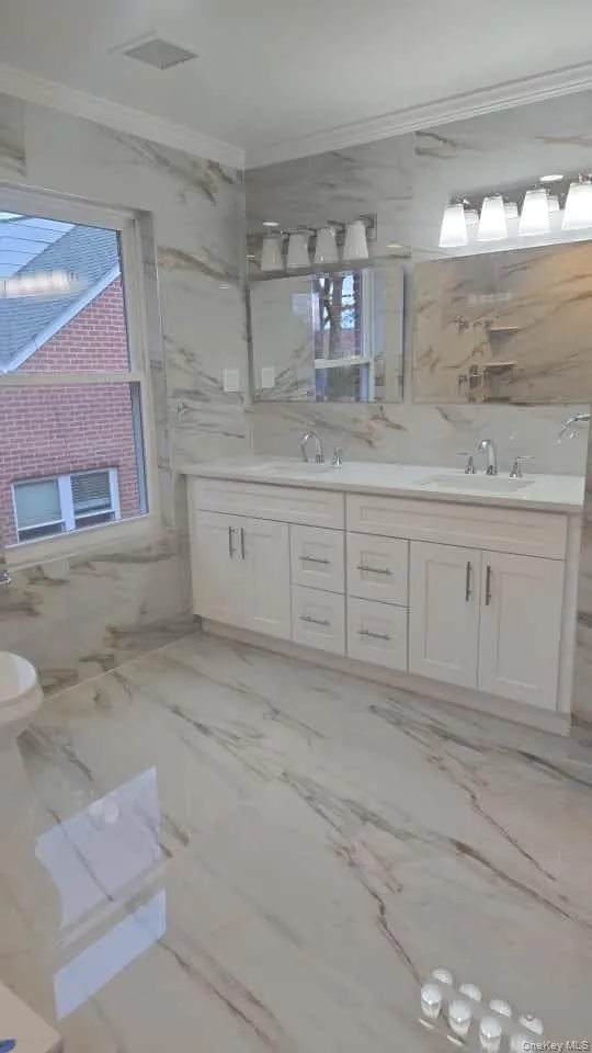 Full bathroom with double vanity, ornamental molding, light marble finish flooring, tile walls, and stone wall Full bathroom with double vanity, ornamental molding, light marble finish flooring, tile walls, and stone wall