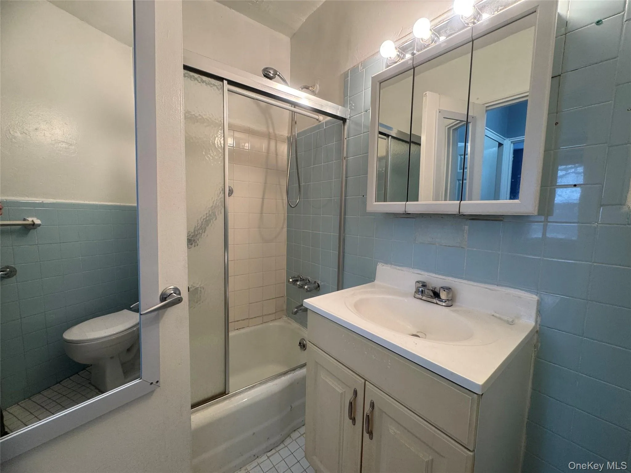 Bathroom featuring tile walls, shower / bath combination with glass door, vanity, a wainscoted wall, and light tile patterned floors Bathroom featuring tile walls, shower / bath combination with glass door, vanity, a wainscoted wall, and light tile patterned floors