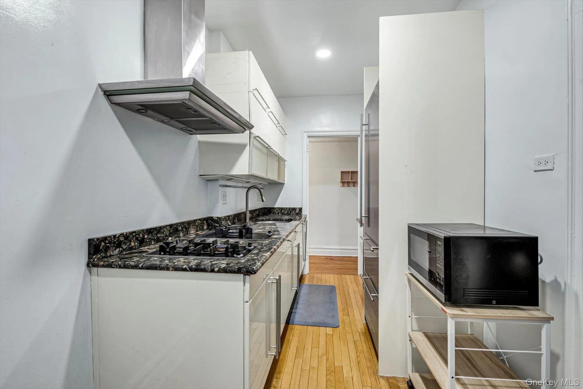 Kitchen with white cabinets, wall chimney range hood, black microwave, dark stone counters, and light wood-type flooring Kitchen with white cabinets, wall chimney range hood, black microwave, dark stone counters, and light wood-type flooring