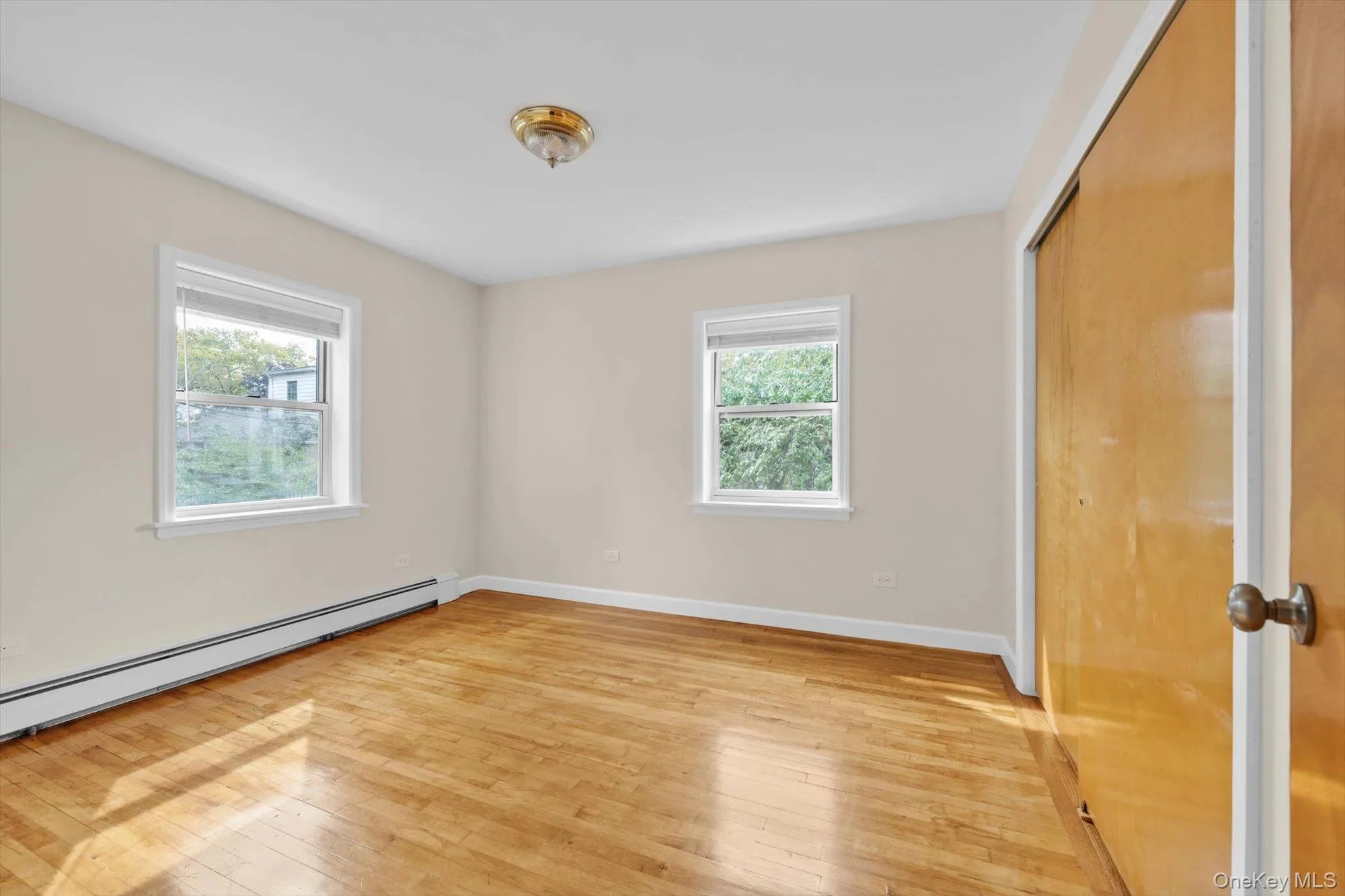 Unfurnished bedroom featuring multiple windows, a closet, light wood-style floors, and baseboard heating Unfurnished bedroom featuring multiple windows, a closet, light wood-style floors, and baseboard heating