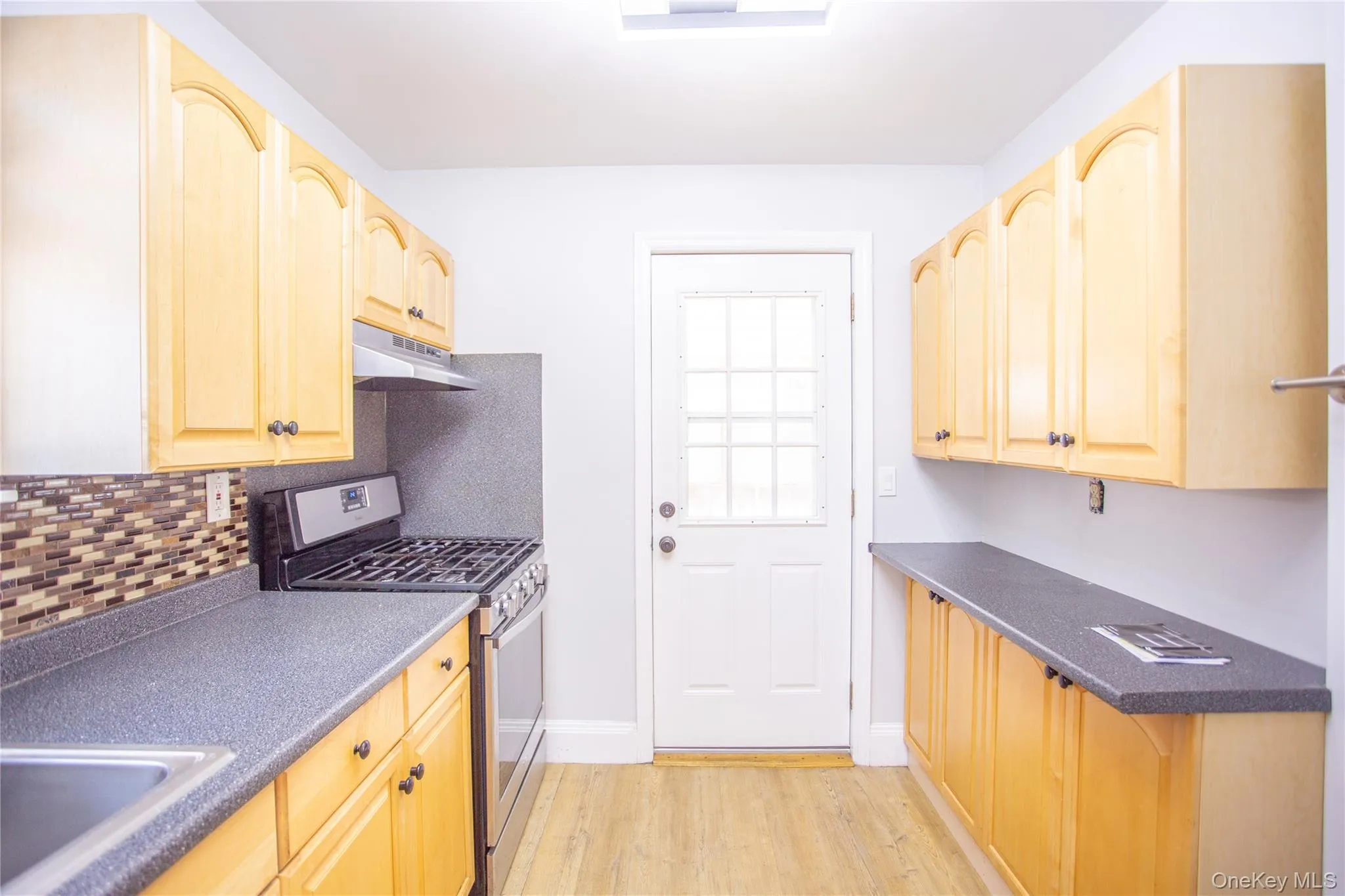 Kitchen featuring light brown cabinetry, gas range, under cabinet range hood, dark countertops, and light wood-style flooring Kitchen featuring light brown cabinetry, gas range, under cabinet range hood, dark countertops, and light wood-style flooring