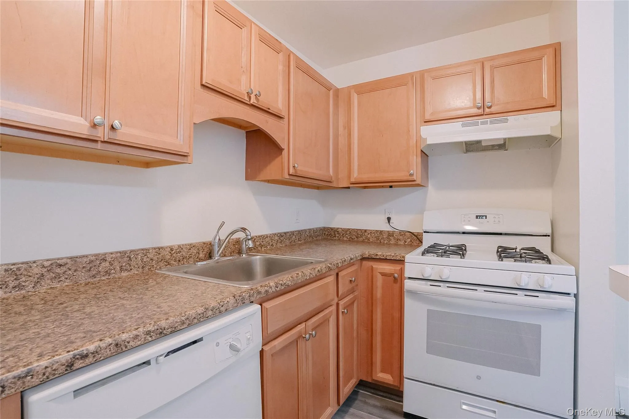 Kitchen with white appliances, light brown cabinetry, under cabinet range hood, and light countertops Kitchen with white appliances, light brown cabinetry, under cabinet range hood, and light countertops