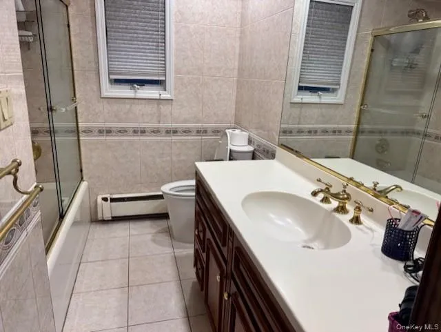 Full bath featuring shower / bath combination with glass door, vanity, light tile patterned floors, tile walls, and a baseboard radiator Full bath featuring shower / bath combination with glass door, vanity, light tile patterned floors, tile walls, and a baseboard radiator