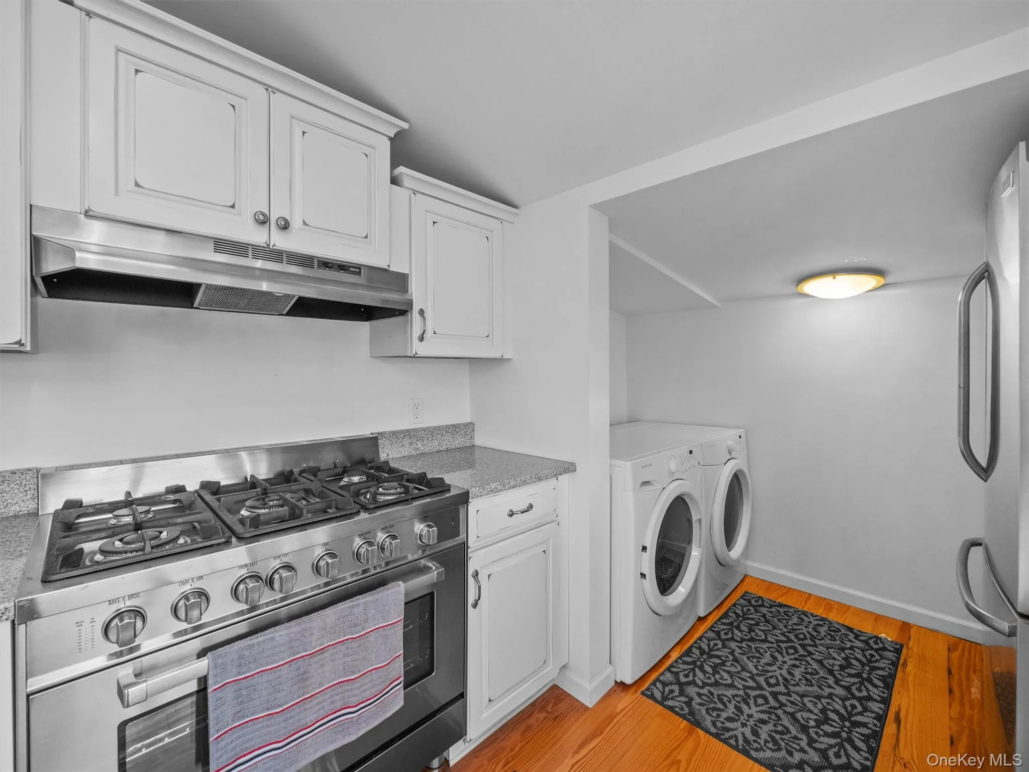 Kitchen featuring appliances with stainless steel finishes, white cabinetry, separate washer and dryer, under cabinet range hood, and light wood finished floors Kitchen featuring appliances with stainless steel finishes, white cabinetry, separate washer and dryer, under cabinet range hood, and light wood finished floors