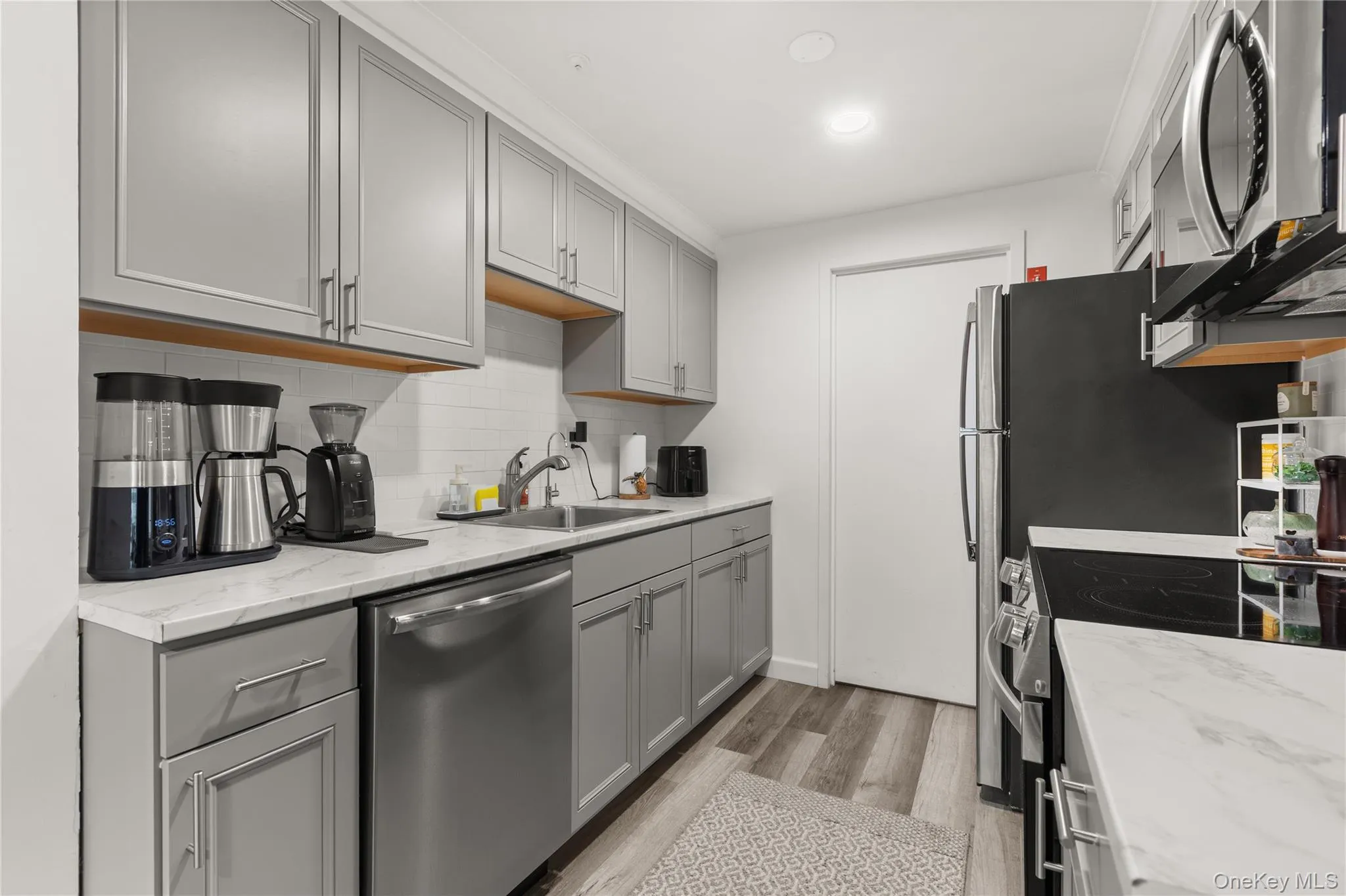 Kitchen with gray cabinets, stainless steel appliances, light wood-style flooring, backsplash, and light stone countertops Kitchen with gray cabinets, stainless steel appliances, light wood-style flooring, backsplash, and light stone countertops