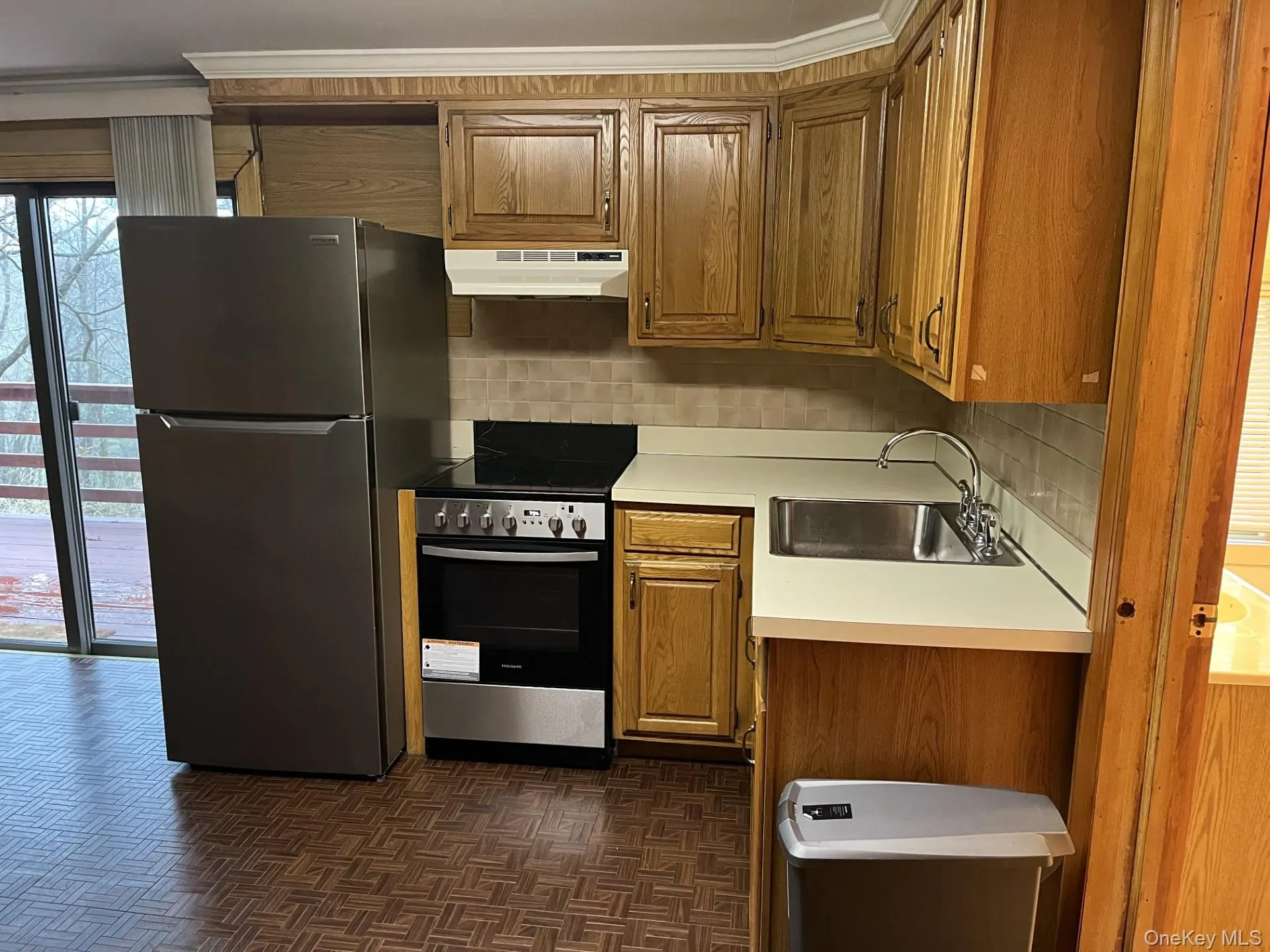 Kitchen featuring stainless steel appliances, light countertops, brown cabinets, under cabinet range hood, and backsplash Kitchen featuring stainless steel appliances, light countertops, brown cabinets, under cabinet range hood, and backsplash