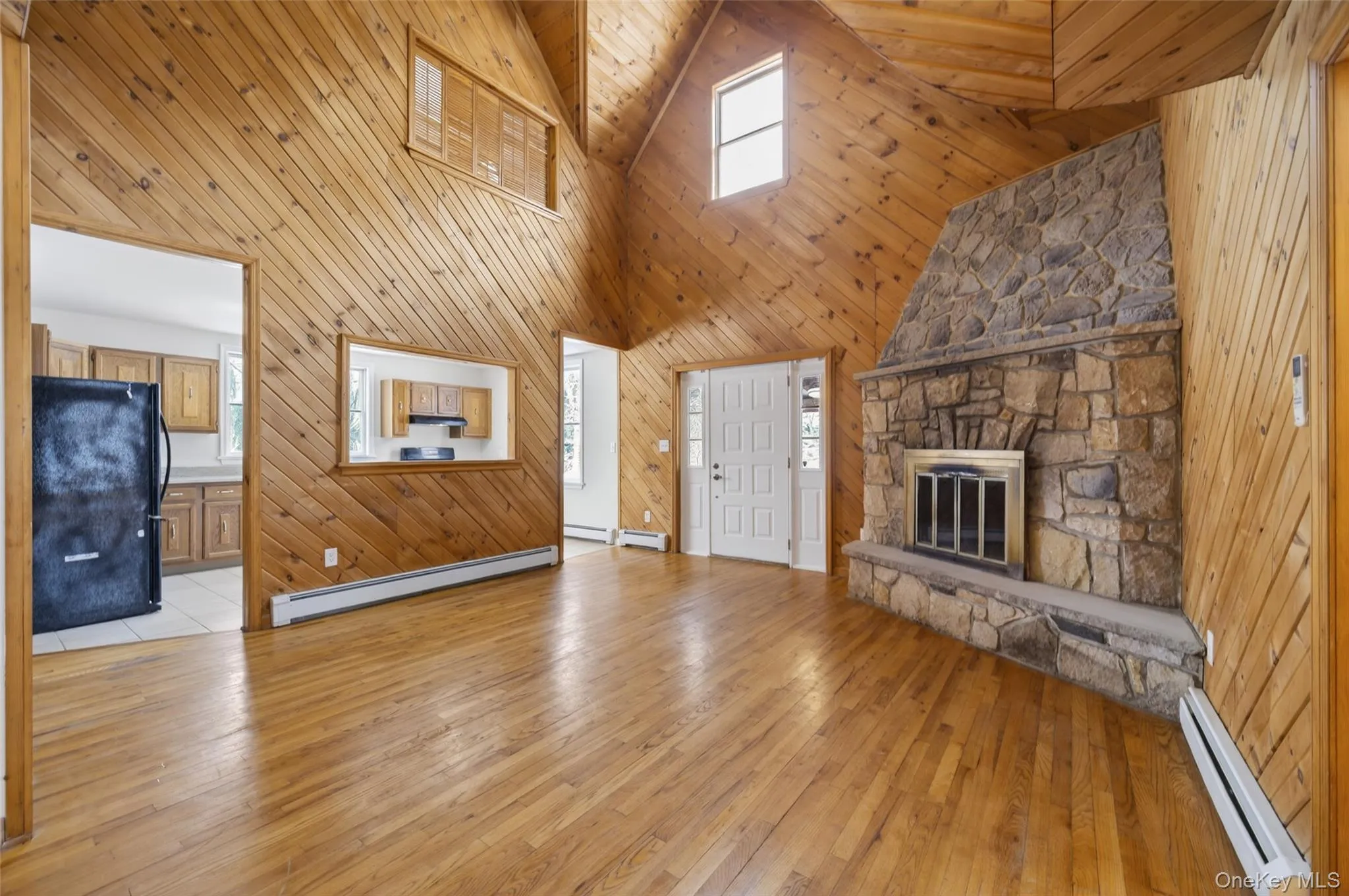 Unfurnished living room featuring wooden walls, light wood-type flooring, baseboard heating, a fireplace, and high vaulted ceiling Unfurnished living room featuring wooden walls, light wood-type flooring, baseboard heating, a fireplace, and high vaulted ceiling