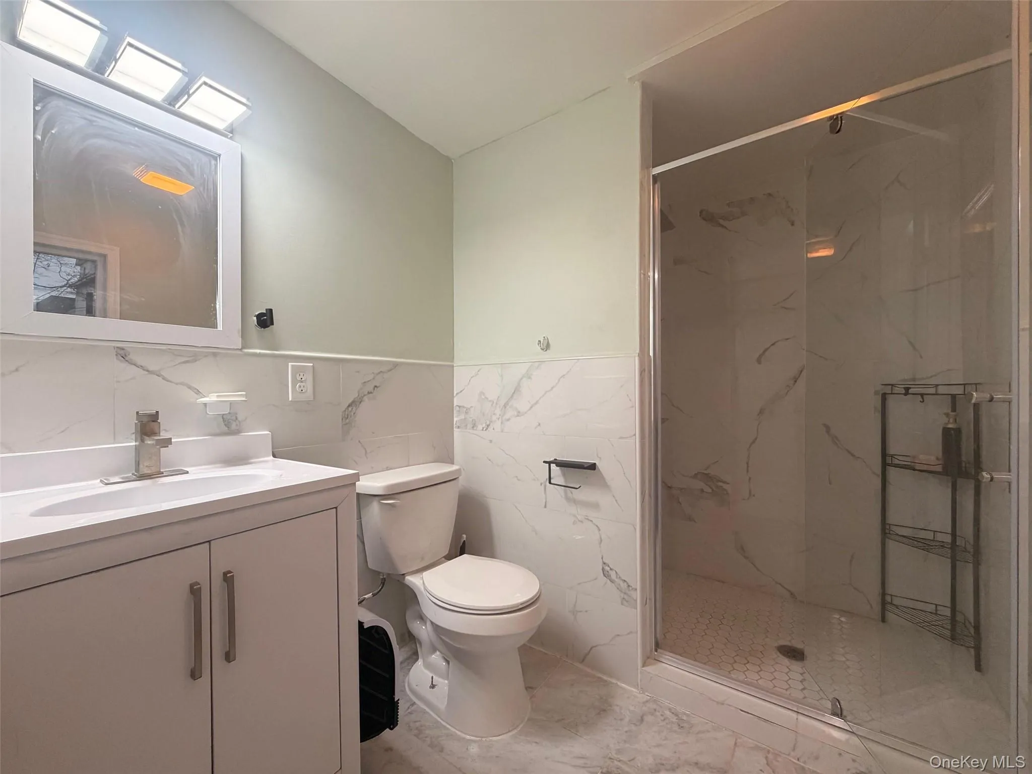 Full bath with a marble finish shower, vanity, tile walls, light marble finish flooring, and wainscoting Full bath with a marble finish shower, vanity, tile walls, light marble finish flooring, and wainscoting