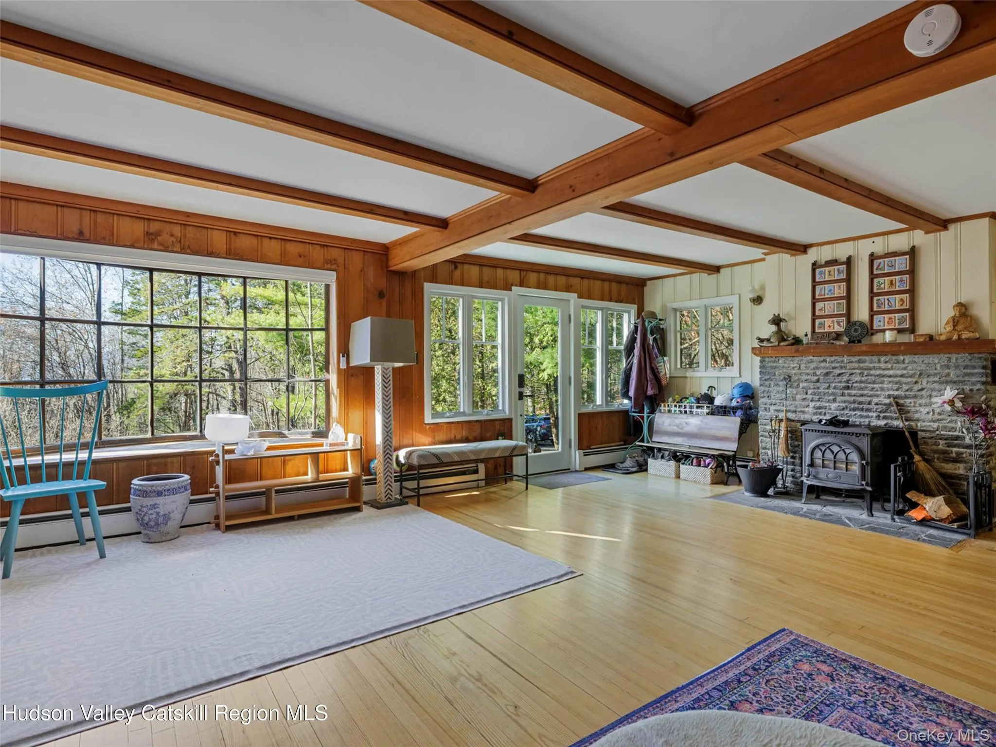 Living area featuring a wood stove, wooden walls, wood-type flooring, beam ceiling, and a baseboard radiator Living area featuring a wood stove, wooden walls, wood-type flooring, beam ceiling, and a baseboard radiator