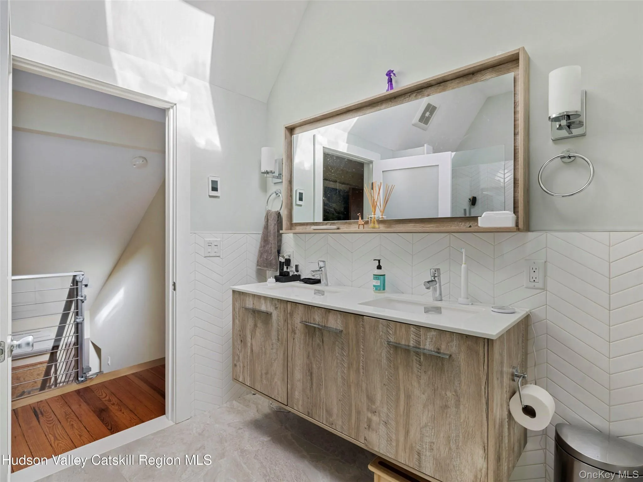 Full bath featuring vaulted ceiling, double vanity, light wood-style flooring, wainscoting, and tile walls Full bath featuring vaulted ceiling, double vanity, light wood-style flooring, wainscoting, and tile walls