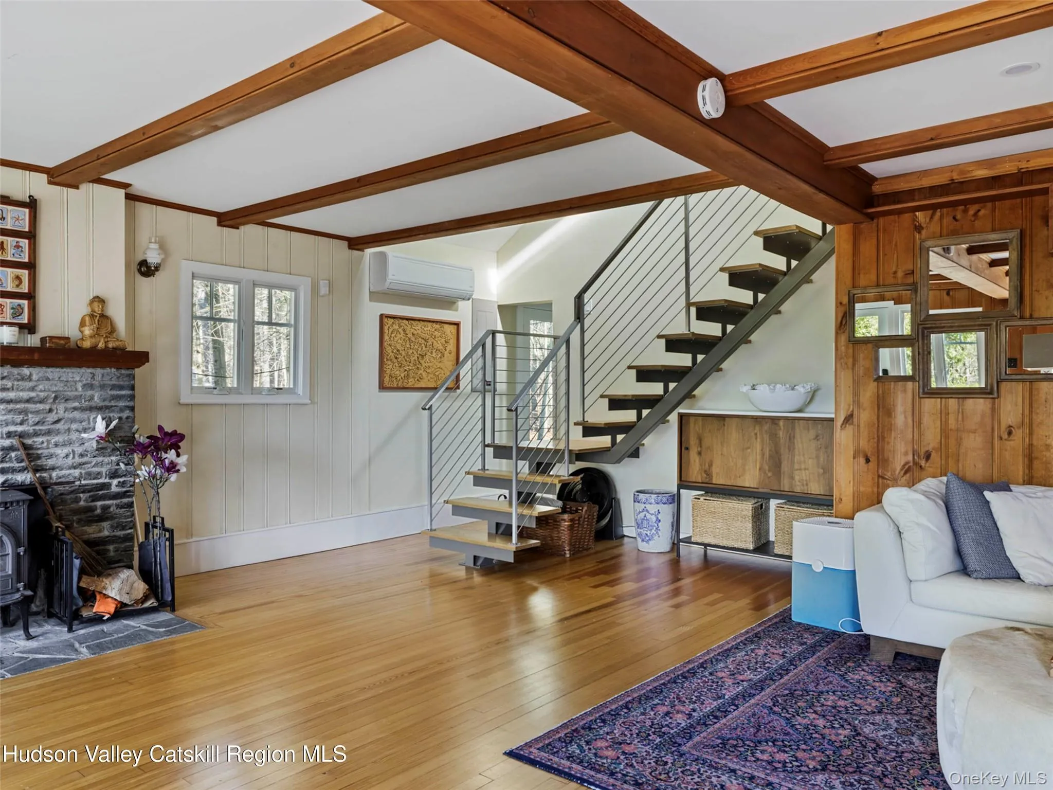 Living room featuring wooden walls, wood-type flooring, beam ceiling, stairs, and a wood stove Living room featuring wooden walls, wood-type flooring, beam ceiling, stairs, and a wood stove