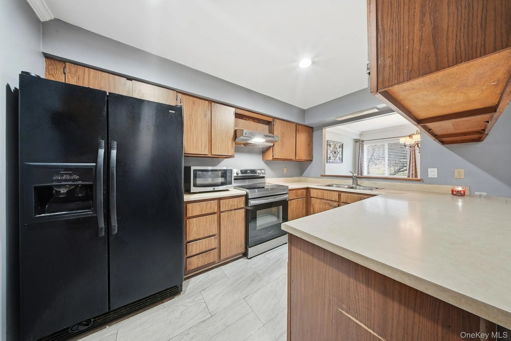 Kitchen featuring appliances with stainless steel finishes, light countertops, range hood, brown cabinetry, and a chandelier Kitchen featuring appliances with stainless steel finishes, light countertops, range hood, brown cabinetry, and a chandelier