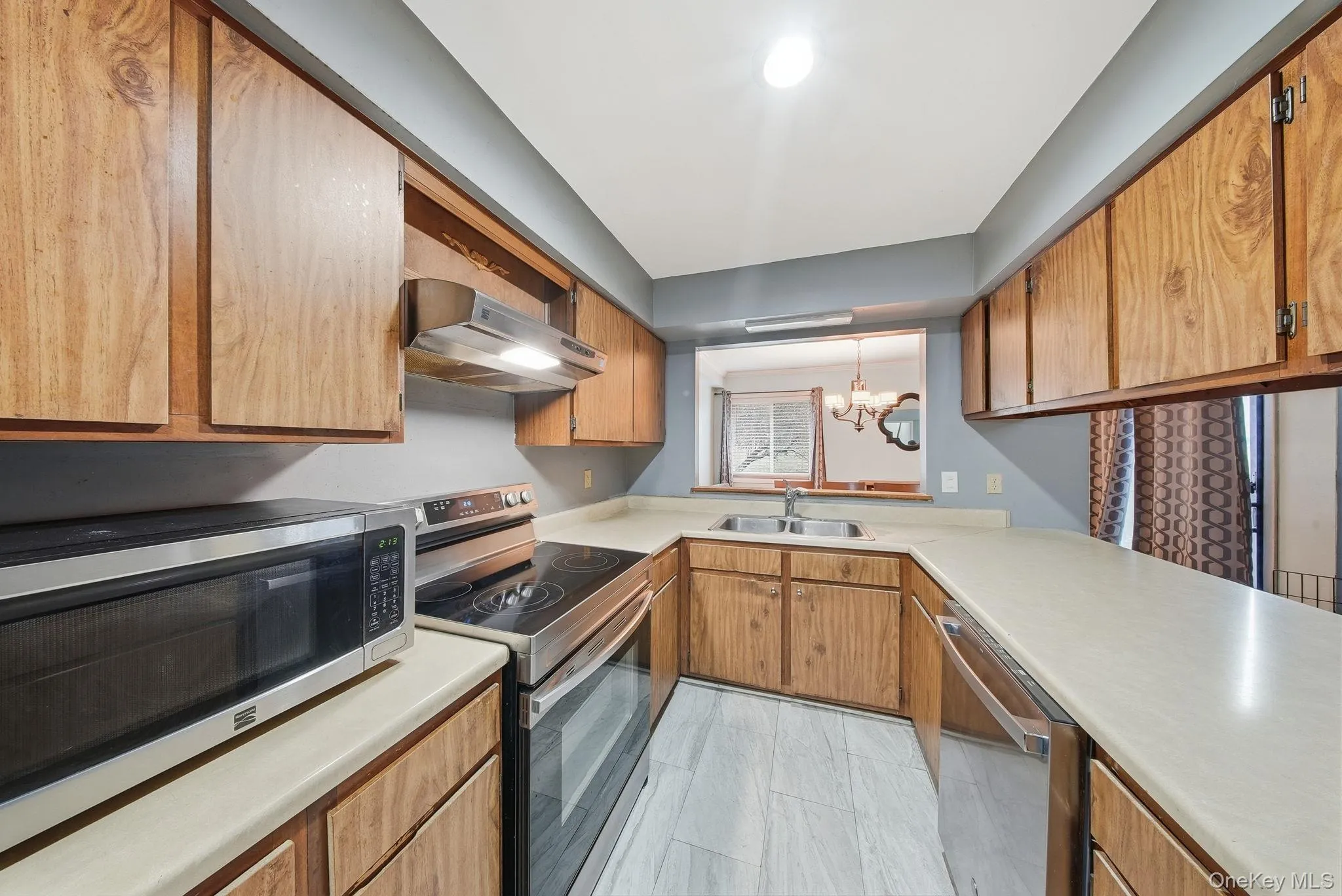 Kitchen with appliances with stainless steel finishes, a chandelier, light countertops, range hood, and brown cabinetry Kitchen with appliances with stainless steel finishes, a chandelier, light countertops, range hood, and brown cabinetry