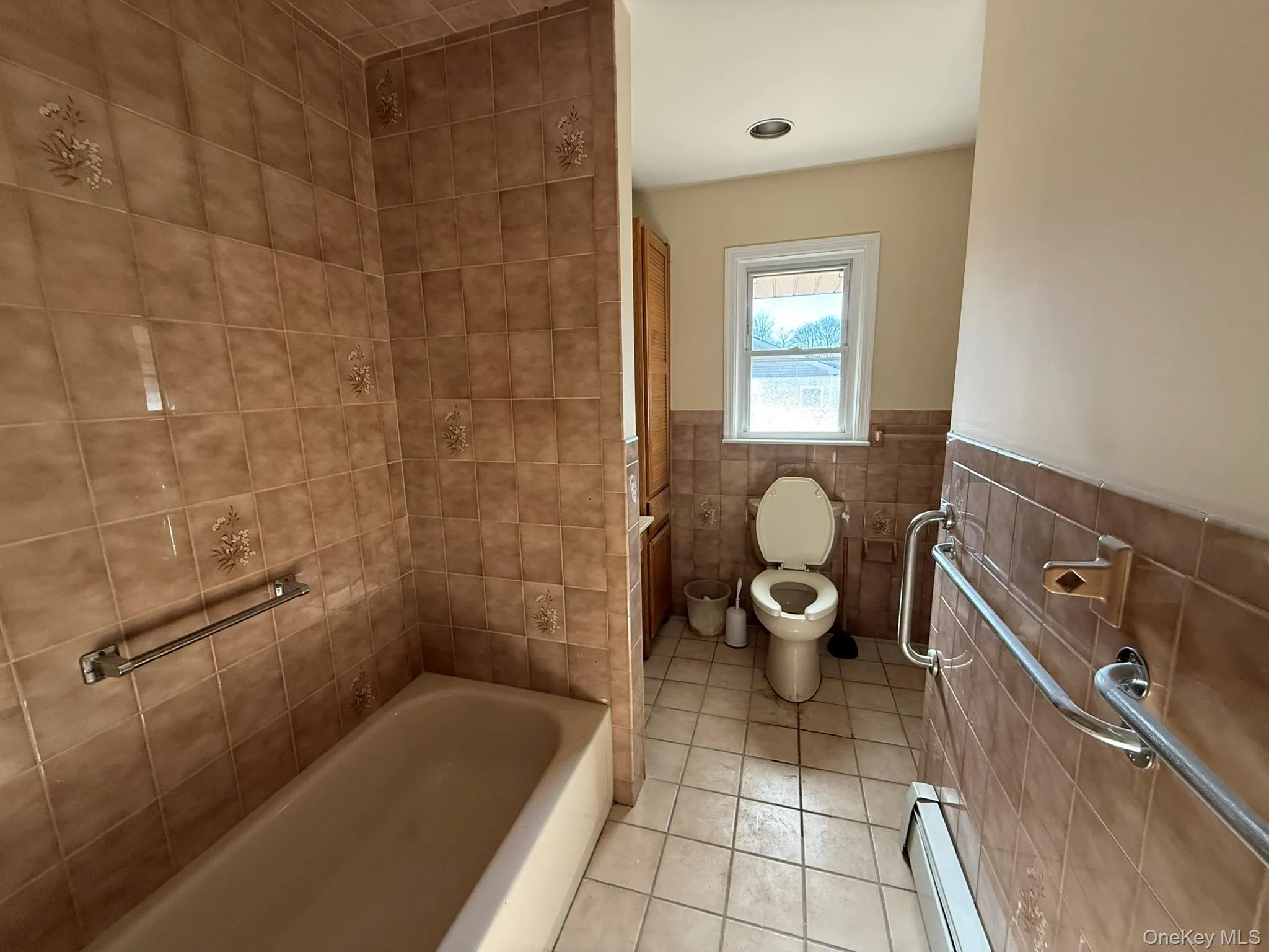 Bathroom featuring light tile patterned flooring, baseboard heating, a tub to relax in, tile walls, and wainscoting Bathroom featuring light tile patterned flooring, baseboard heating, a tub to relax in, tile walls, and wainscoting