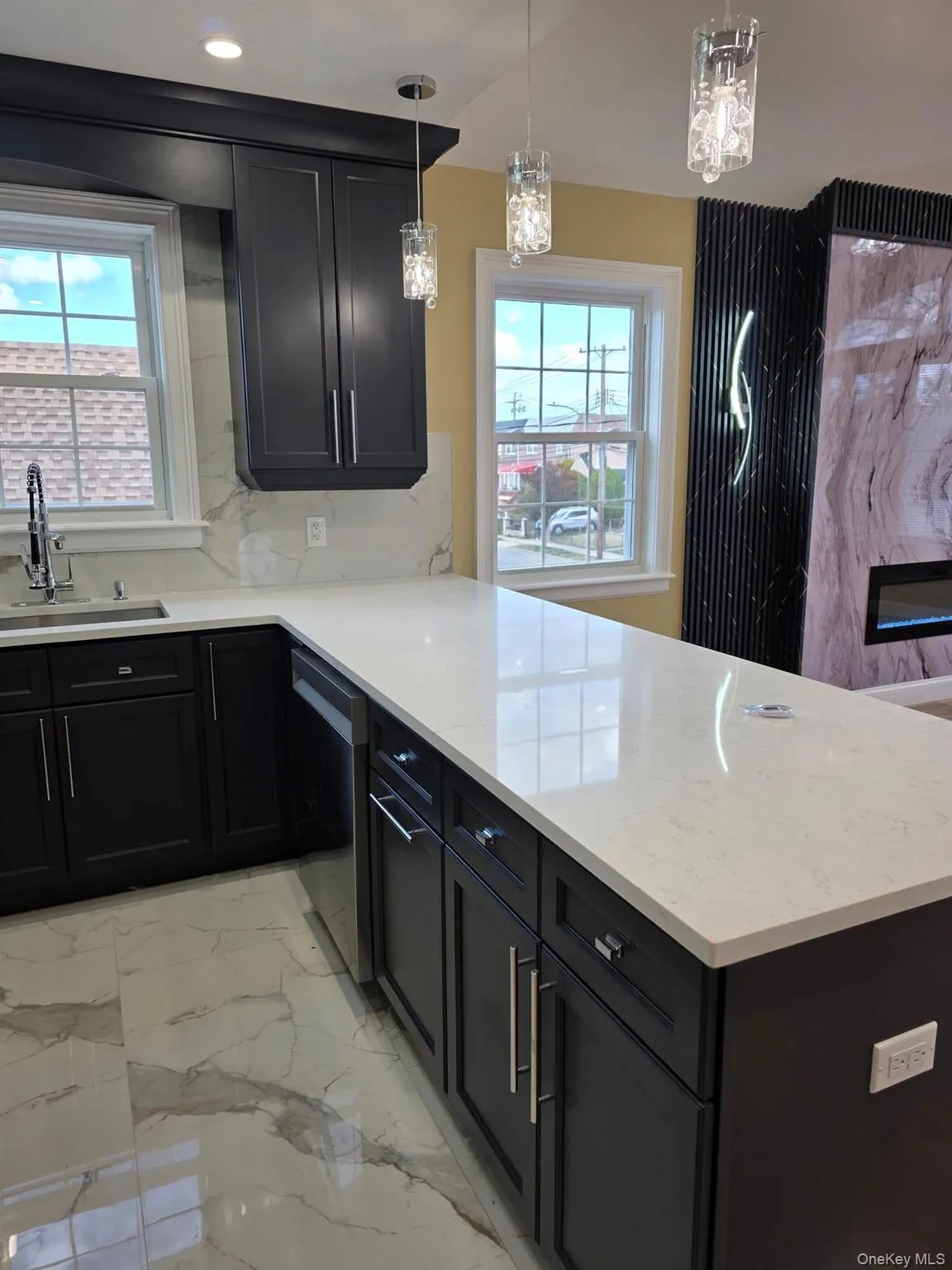 Kitchen with decorative light fixtures, decorative backsplash, dark cabinetry, a peninsula, and light marble finish flooring Kitchen with decorative light fixtures, decorative backsplash, dark cabinetry, a peninsula, and light marble finish flooring