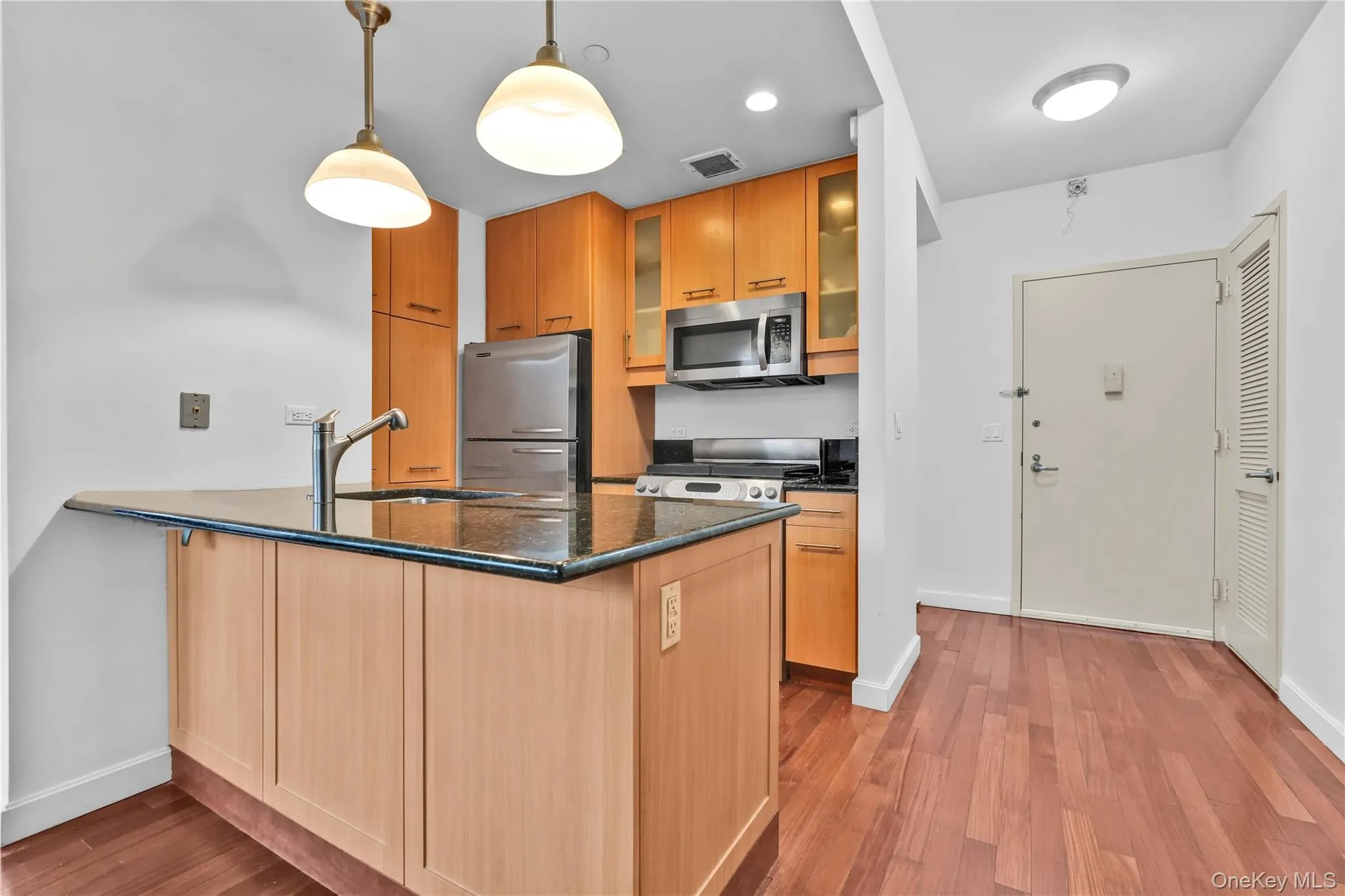 Kitchen featuring dark stone counters, appliances with stainless steel finishes, a peninsula, glass insert cabinets, and hanging light fixtures Kitchen featuring dark stone counters, appliances with stainless steel finishes, a peninsula, glass insert cabinets, and hanging light fixtures