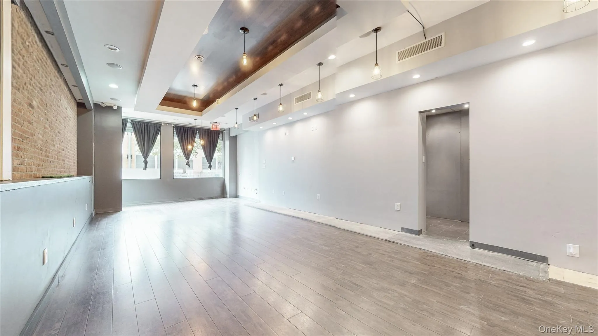 Empty room with a raised ceiling, brick wall, light wood-type flooring, and recessed lighting Empty room with a raised ceiling, brick wall, light wood-type flooring, and recessed lighting