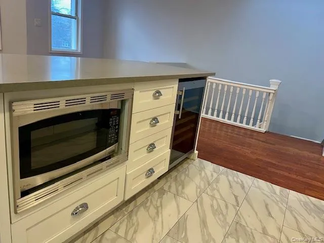 Kitchen featuring stainless steel microwave, light marble finish floors, white cabinets, wine cooler, and light stone countertops Kitchen featuring stainless steel microwave, light marble finish floors, white cabinets, wine cooler, and light stone countertops