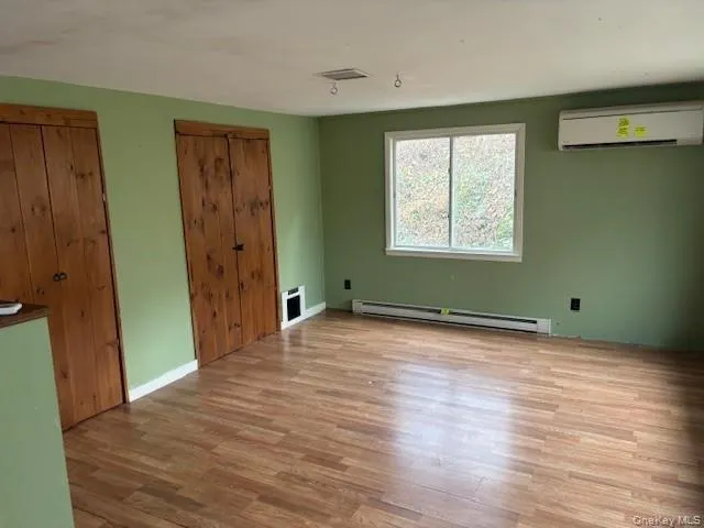 Unfurnished bedroom featuring light wood-type flooring, two closets, baseboard heating, and a wall unit AC Unfurnished bedroom featuring light wood-type flooring, two closets, baseboard heating, and a wall unit AC