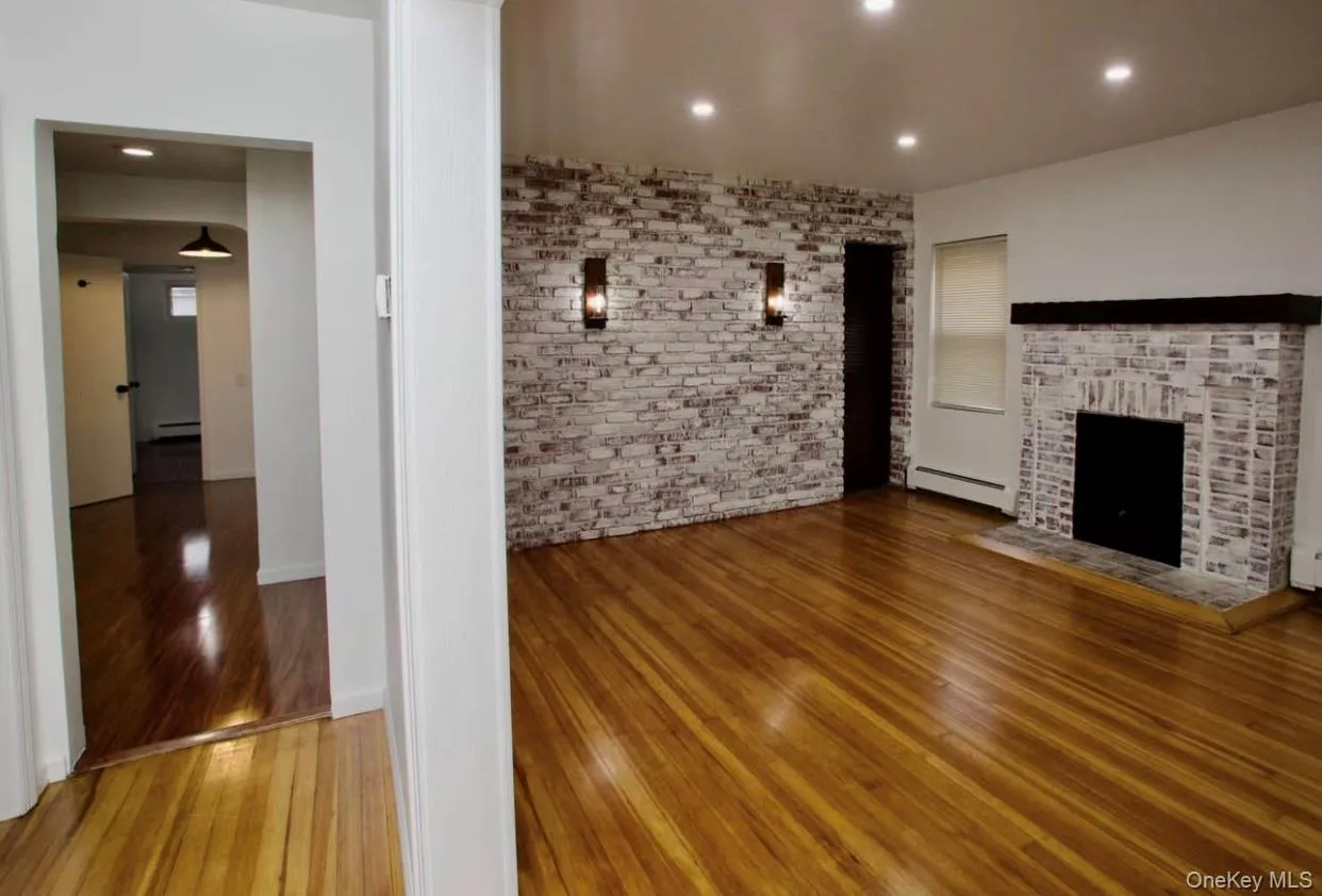 Unfurnished living room featuring wood-type flooring, recessed lighting, a fireplace, a baseboard heating unit, and brick wall Unfurnished living room featuring wood-type flooring, recessed lighting, a fireplace, a baseboard heating unit, and brick wall