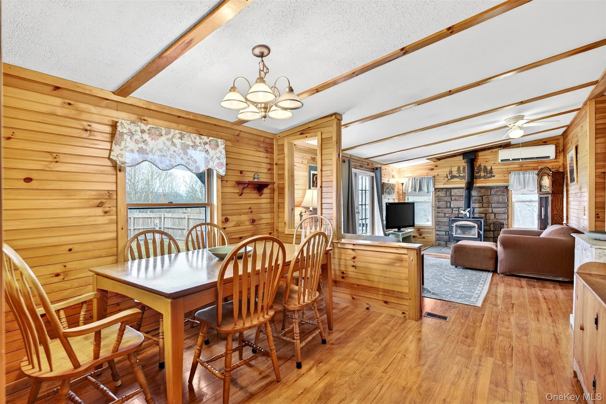 Dining room with a wood stove, beam ceiling, wooden walls, light wood-style floors, and a chandelier Dining room with a wood stove, beam ceiling, wooden walls, light wood-style floors, and a chandelier