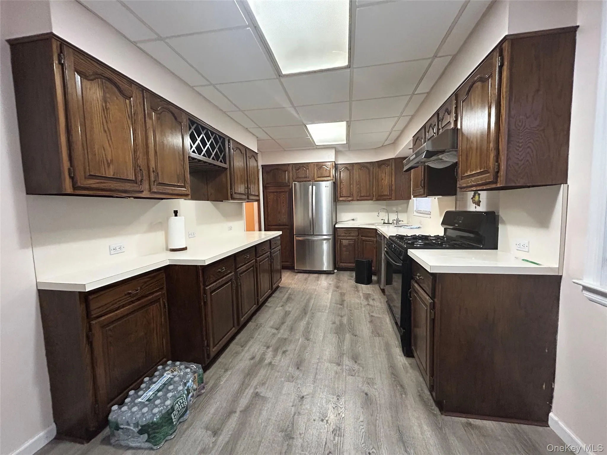Kitchen with light countertops, black gas range oven, dark brown cabinets, freestanding refrigerator, and a paneled ceiling Kitchen with light countertops, black gas range oven, dark brown cabinets, freestanding refrigerator, and a paneled ceiling