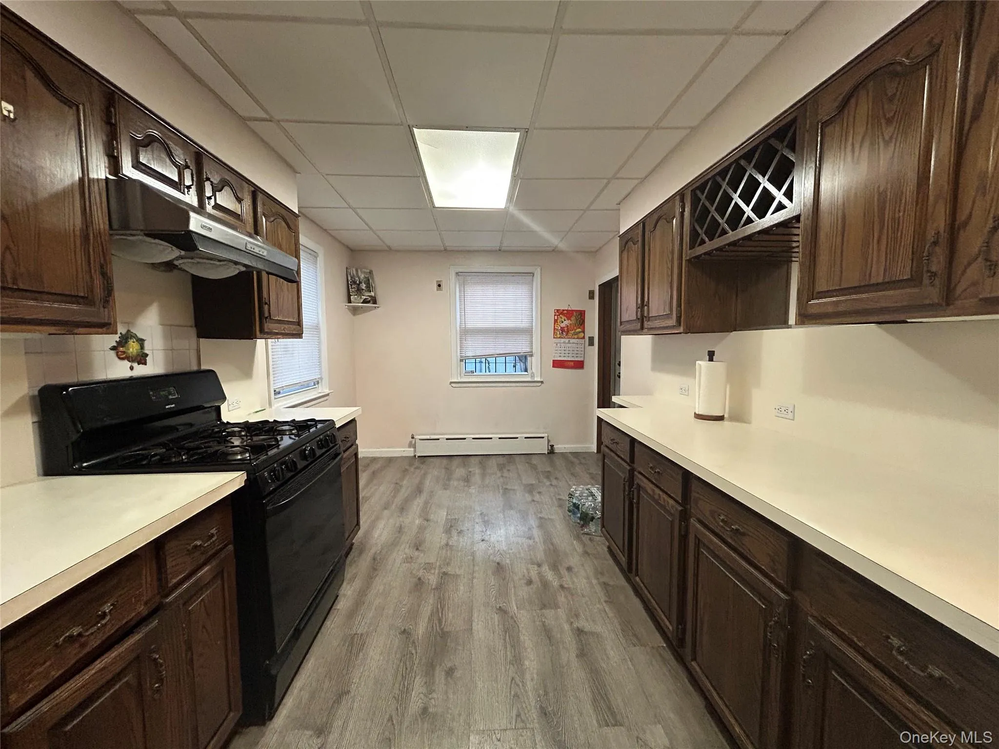 Kitchen with dark brown cabinetry, black range with gas cooktop, under cabinet range hood, light countertops, and a paneled ceiling Kitchen with dark brown cabinetry, black range with gas cooktop, under cabinet range hood, light countertops, and a paneled ceiling