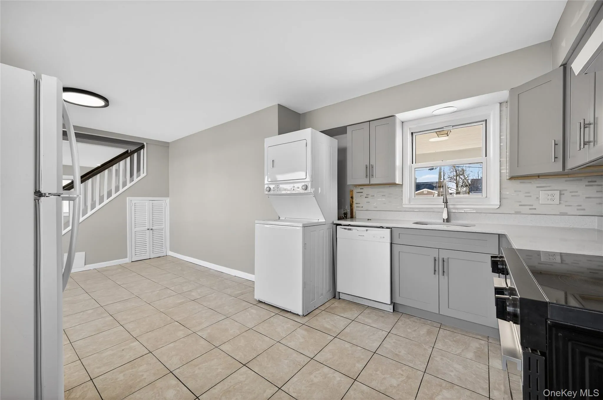 Kitchen featuring gray cabinets, white appliances, decorative backsplash, light tile patterned floors, and stacked washer and clothes dryer Kitchen featuring gray cabinets, white appliances, decorative backsplash, light tile patterned floors, and stacked washer and clothes dryer