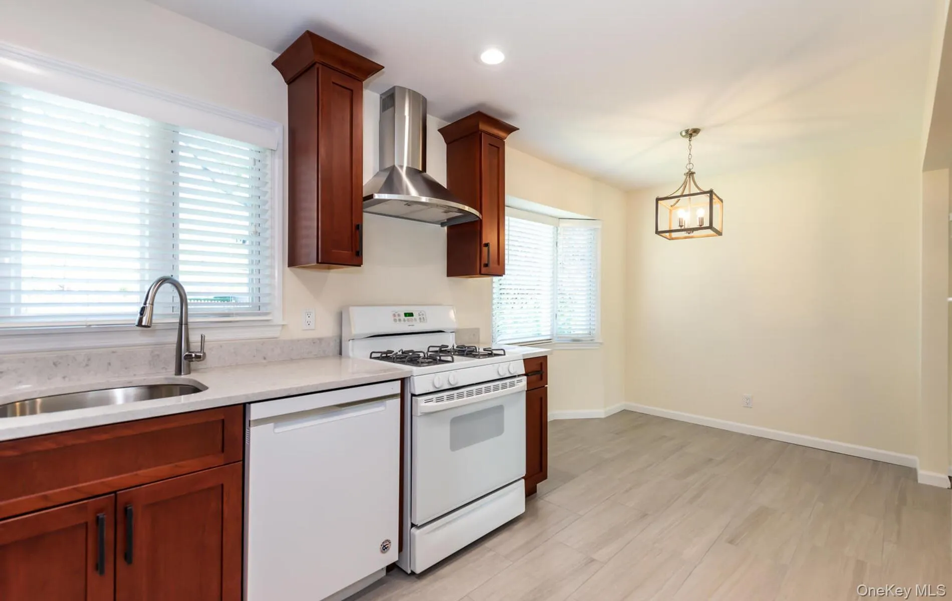 Kitchen with white appliances, wall chimney exhaust hood, decorative light fixtures, light stone countertops, and brown cabinetry Kitchen with white appliances, wall chimney exhaust hood, decorative light fixtures, light stone countertops, and brown cabinetry