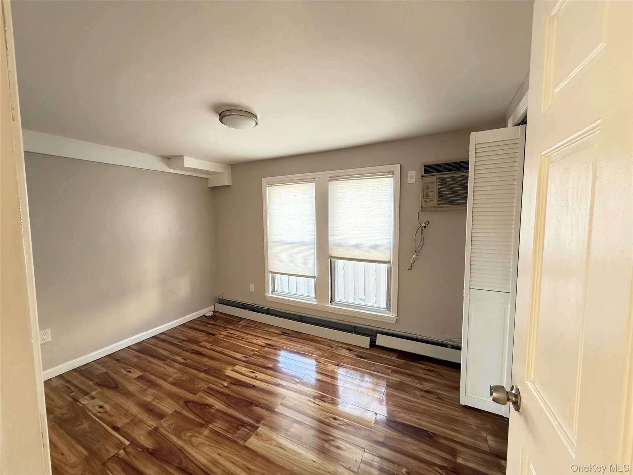 Unfurnished bedroom featuring baseboard heating, dark wood-style floors, and a wall unit AC Unfurnished bedroom featuring baseboard heating, dark wood-style floors, and a wall unit AC